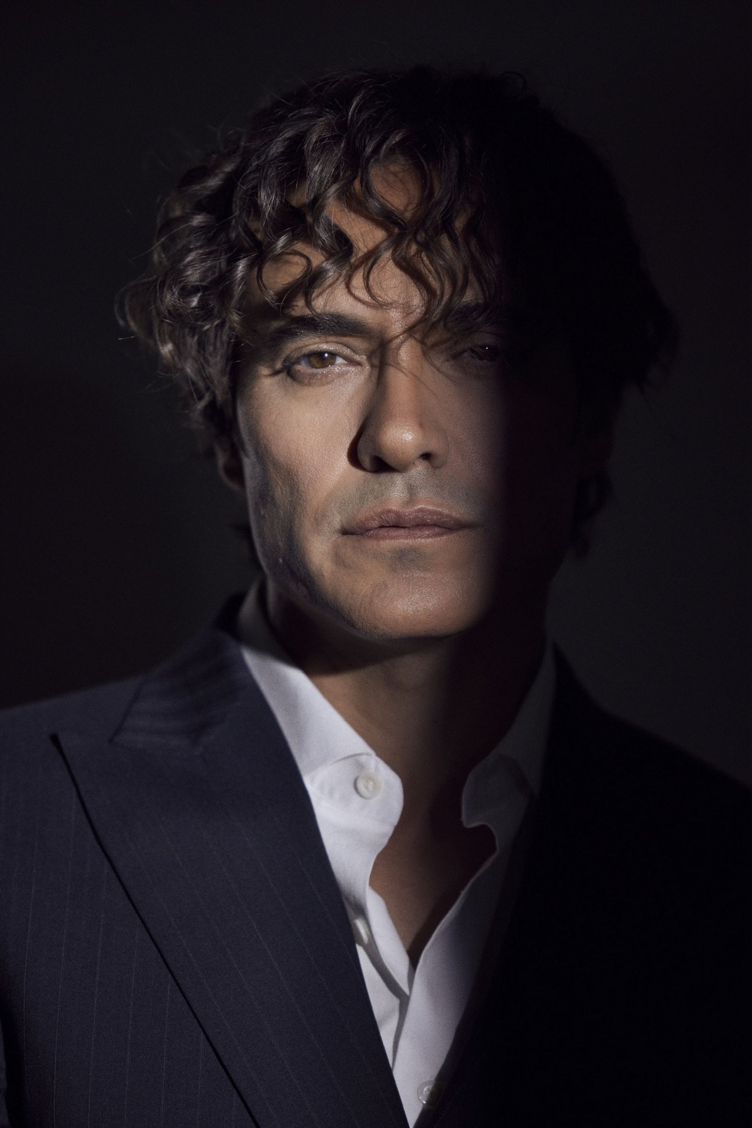 A man with curly brown hair, wearing a dark suit and white shirt, looking directly at the camera with a serious expression against a dark background.