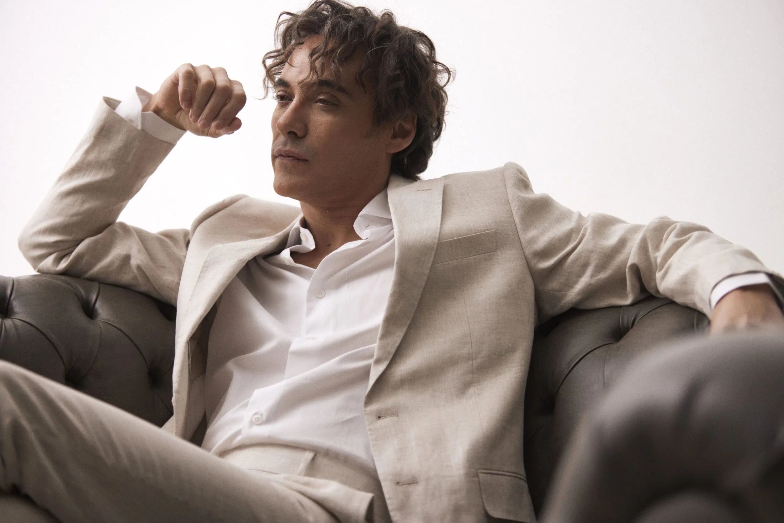 A man in a light-colored suit and white shirt sitting on a leather sofa, flexing his right arm in a confident pose.