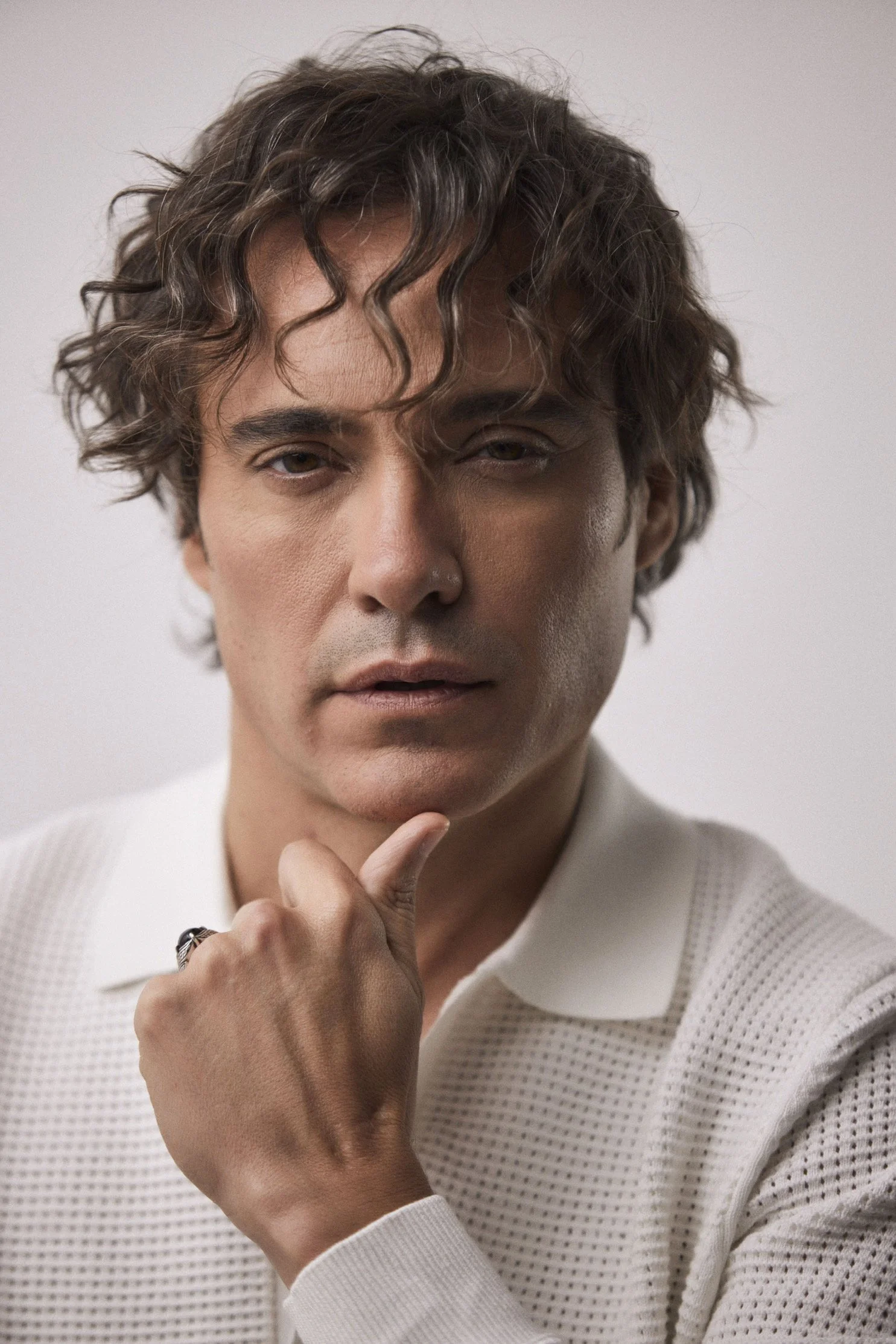 A man with wavy brown hair, light skin, and wearing a white textured shirt, is resting his chin on his thumb with a thoughtful expression against a plain light background.