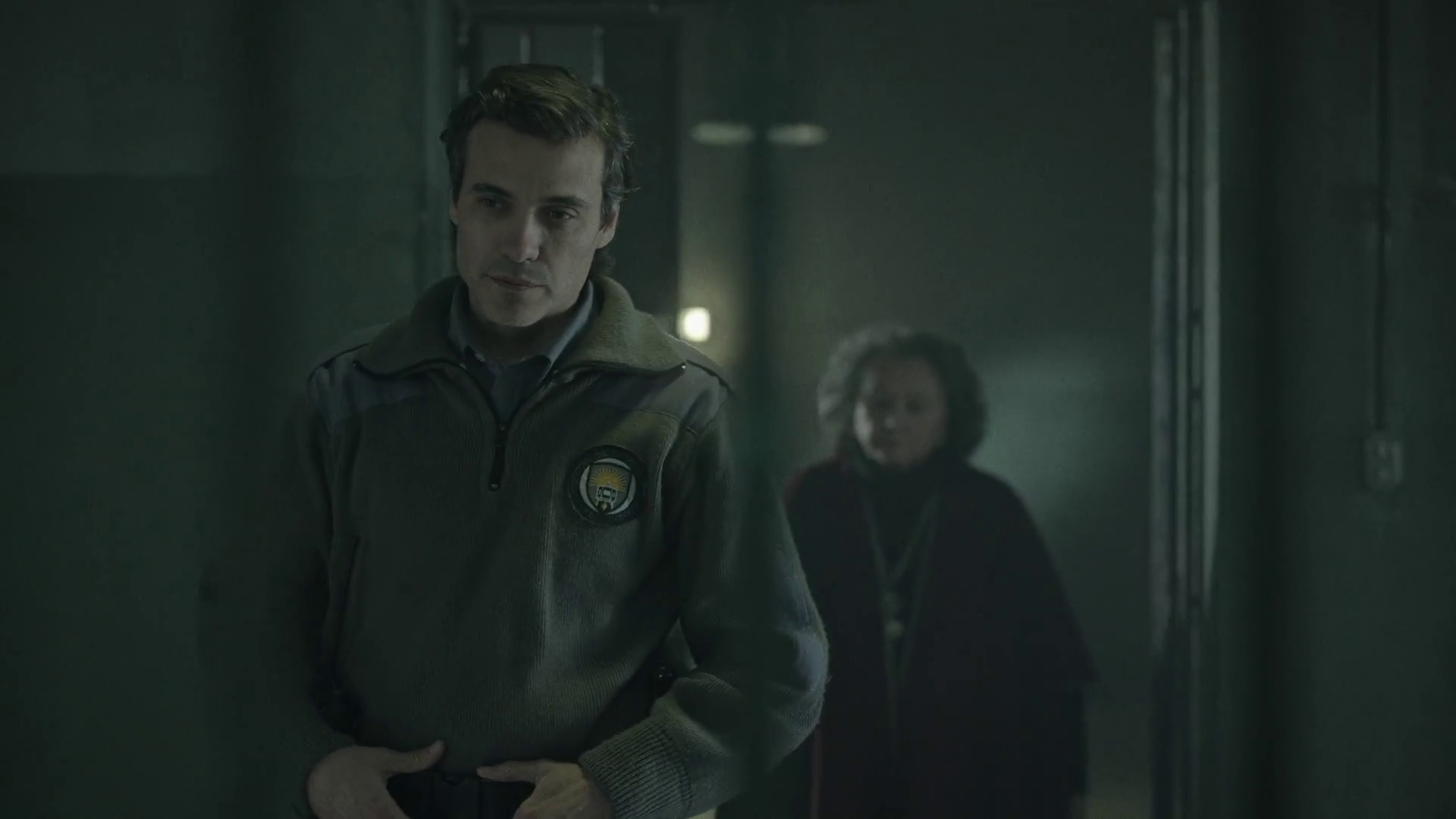 A man in a security or police uniform with a badge on his chest standing in a dimly lit hallway, with a woman in the background.