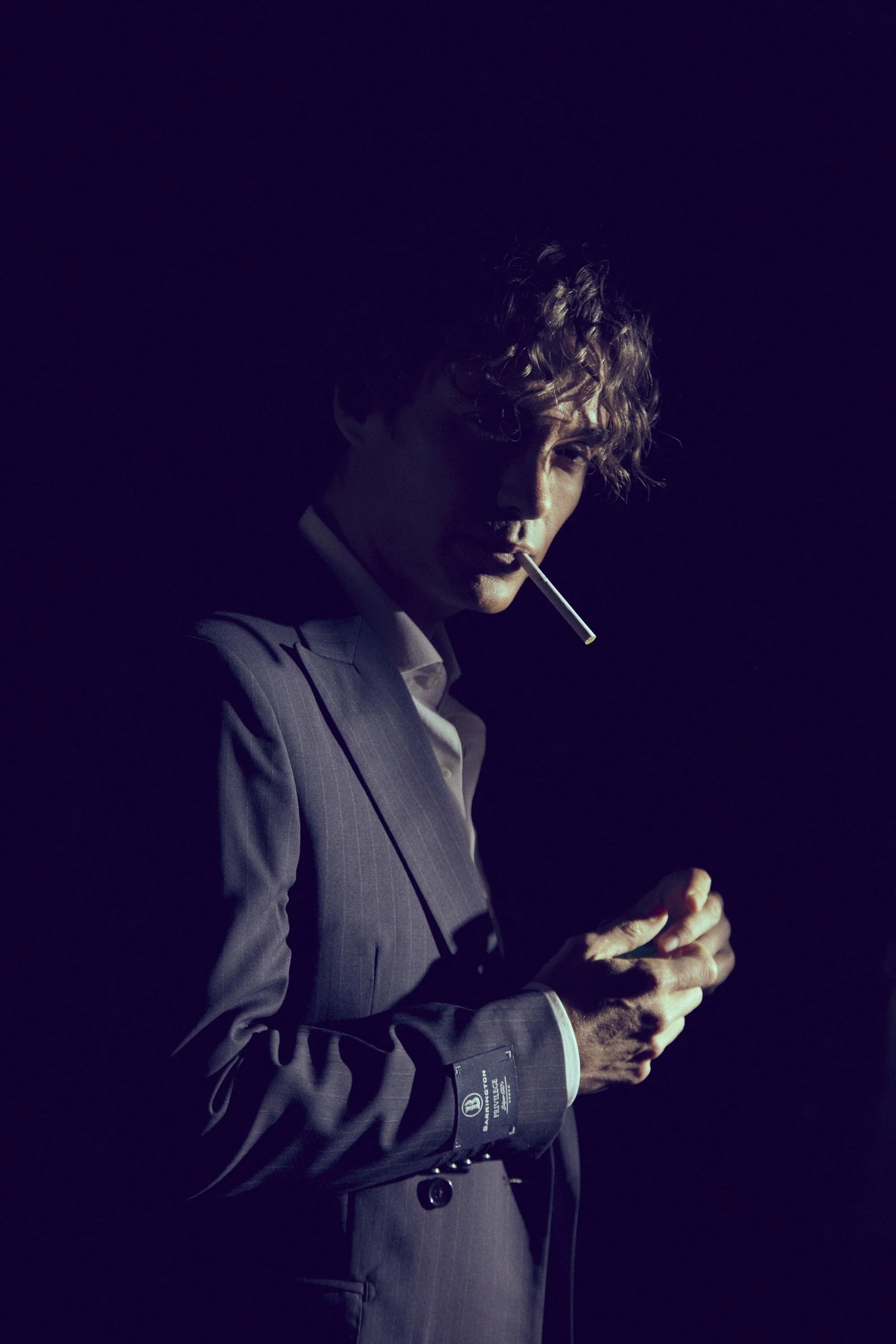 A man in a suit with curly hair, holding a cigarette in his mouth, stands in a dark setting with purple lighting.