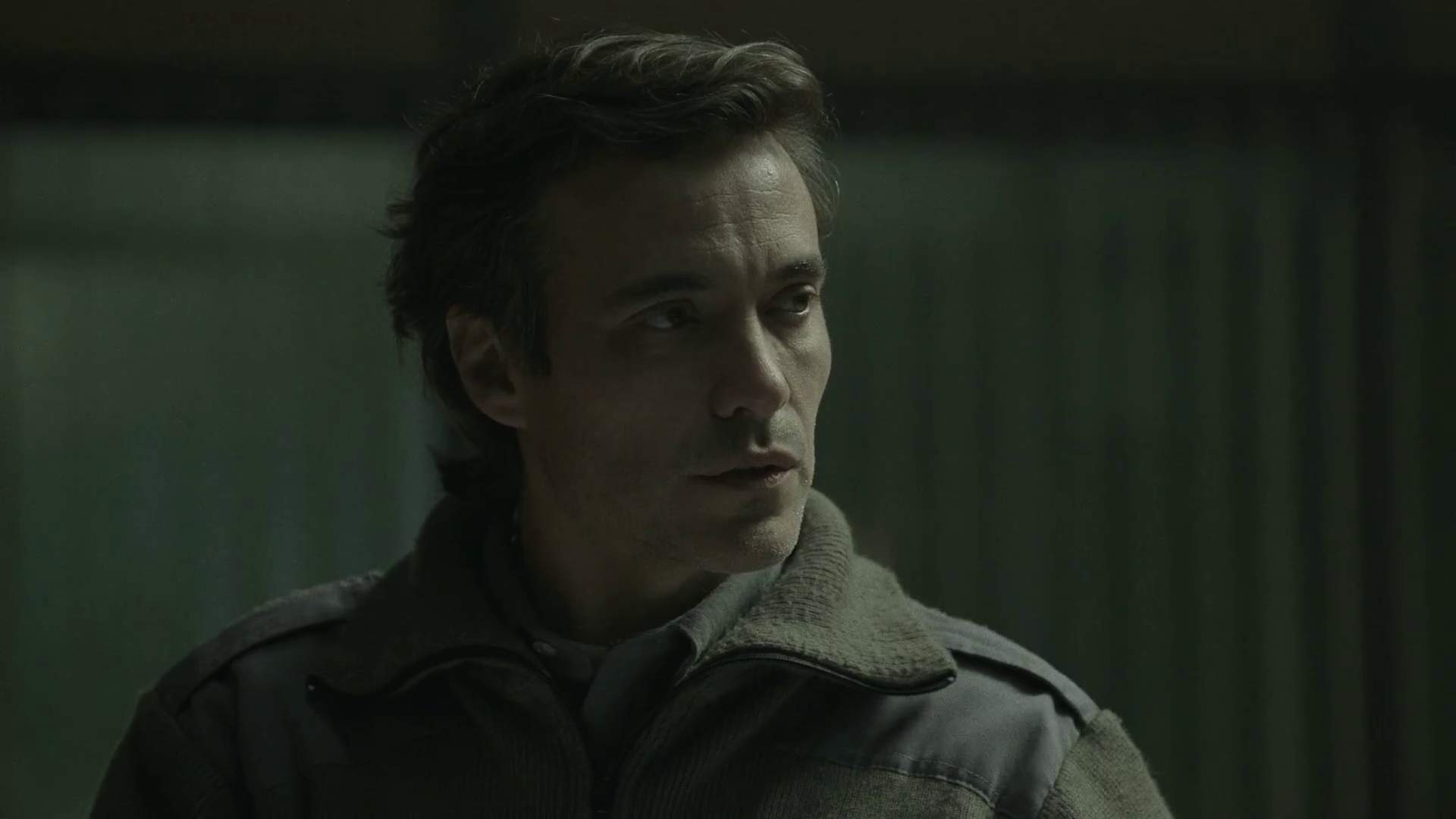 A man with brown hair in a gray jacket looks to the side in a dimly lit room.