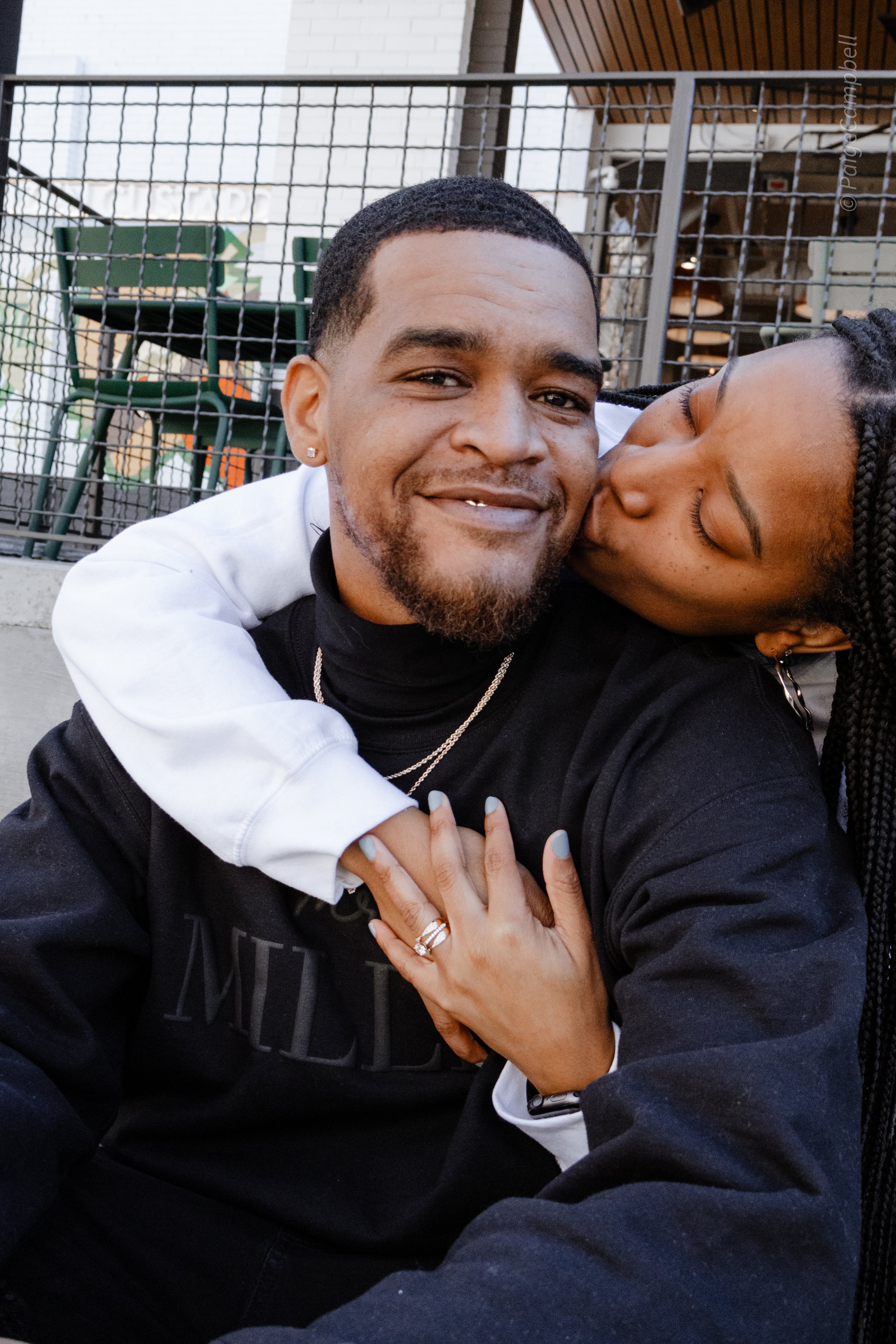 A woman kissing a smiling man's cheek outside; she's embracing him with a ring on her finger.