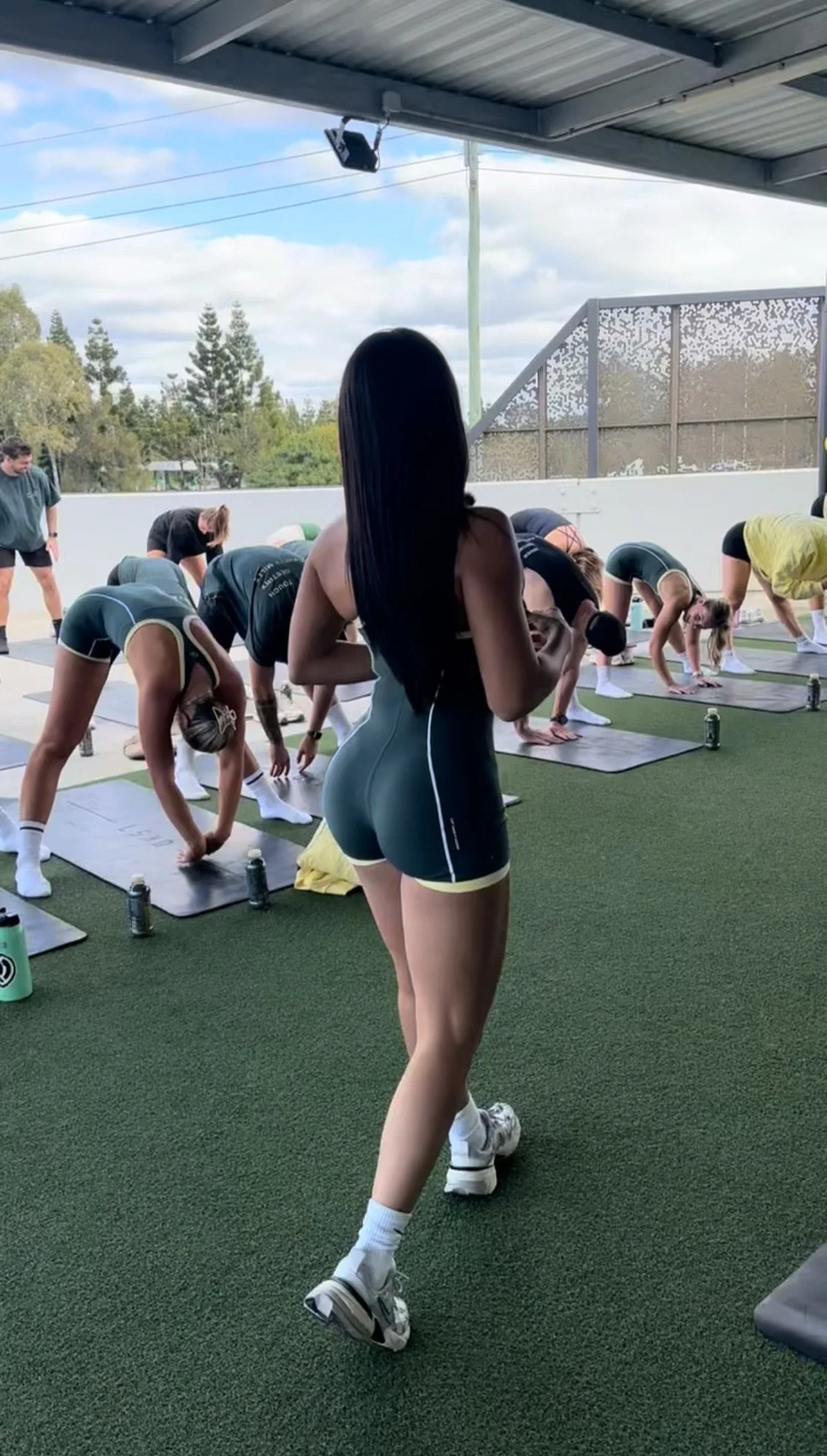 A woman with long black hair in athletic wear is observing a group exercise class at an outdoor fitness area, with trees and a fence in the background.