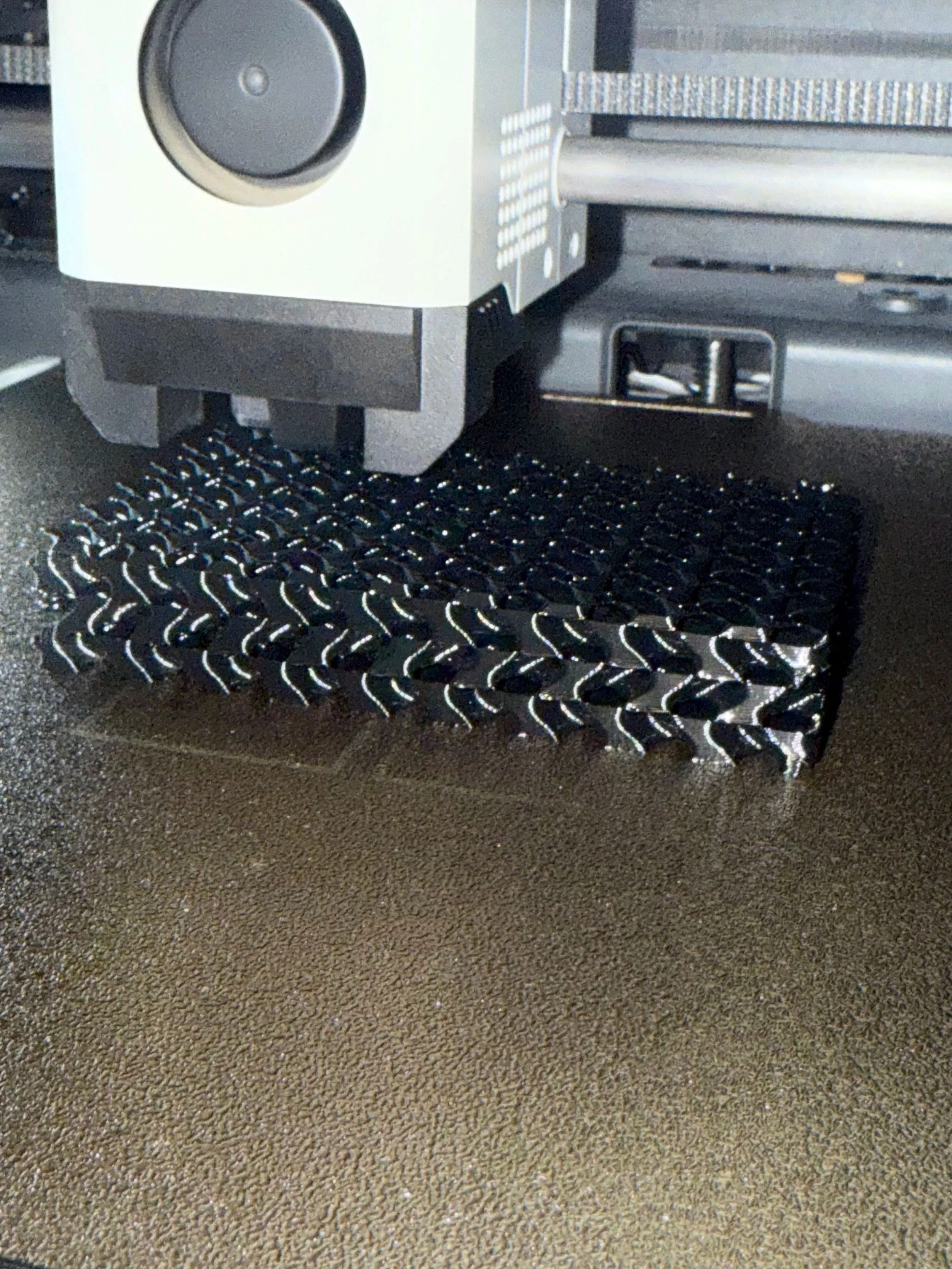 Close-up of a 3D printer nozzle extruding black filament onto a printed object with a lattice pattern.