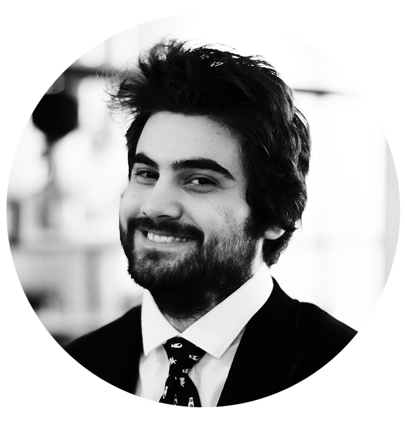 Black and white portrait of Zhoslen Makoev a smiling man with dark, tousled hair and a beard, wearing a suit and tie, in an indoor setting.