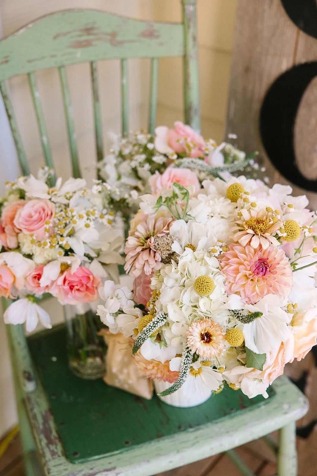 A collection of pastel-colored flowers including roses, dahlias, and daisies arranged in a white vase on a green wooden chair with peeling paint.