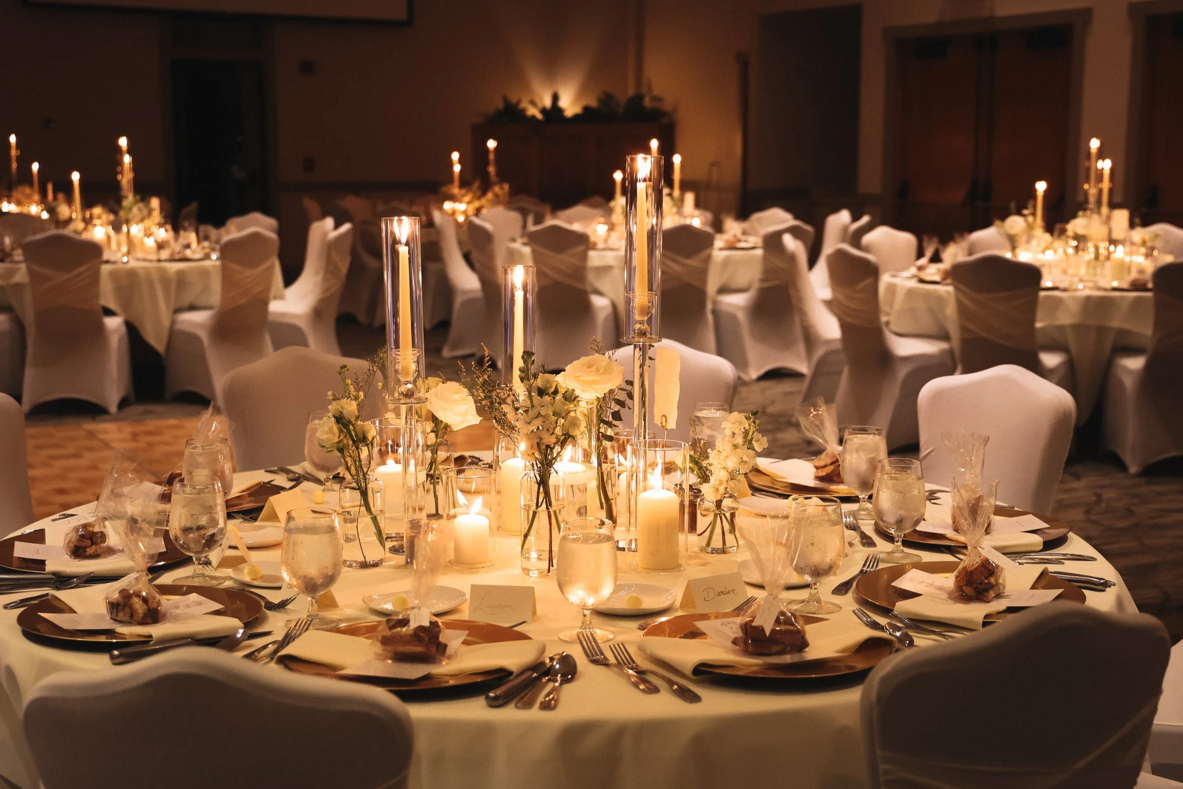 Round banquet table decorated with white floral centerpieces, candles, and tableware, set for an event in a dimly lit banquet hall.