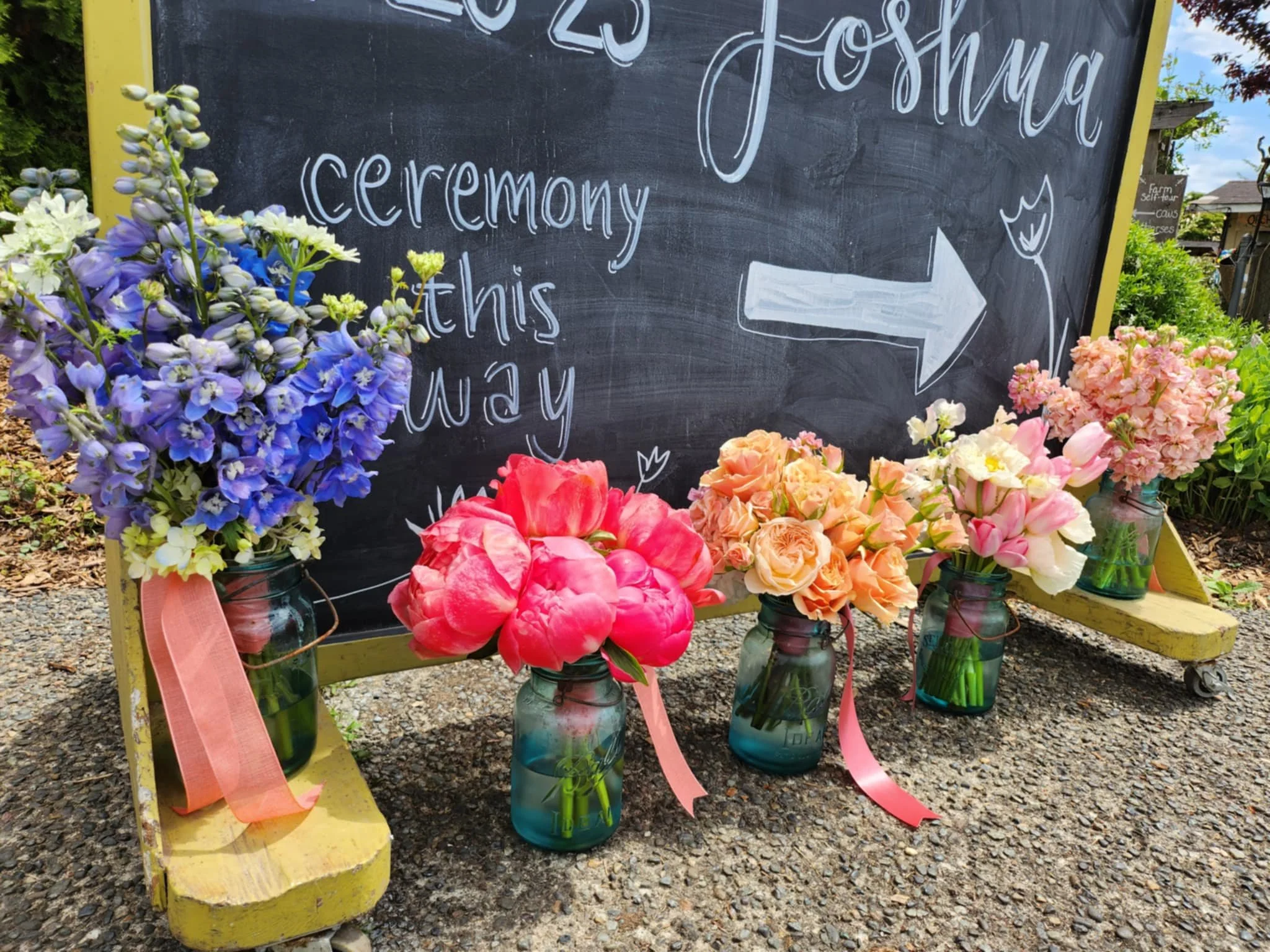 A chalkboard sign with a yellow frame and wheels, decorated with colorful flower arrangements in glass jars, indicating a ceremony at Joshua with an arrow pointing to the right.