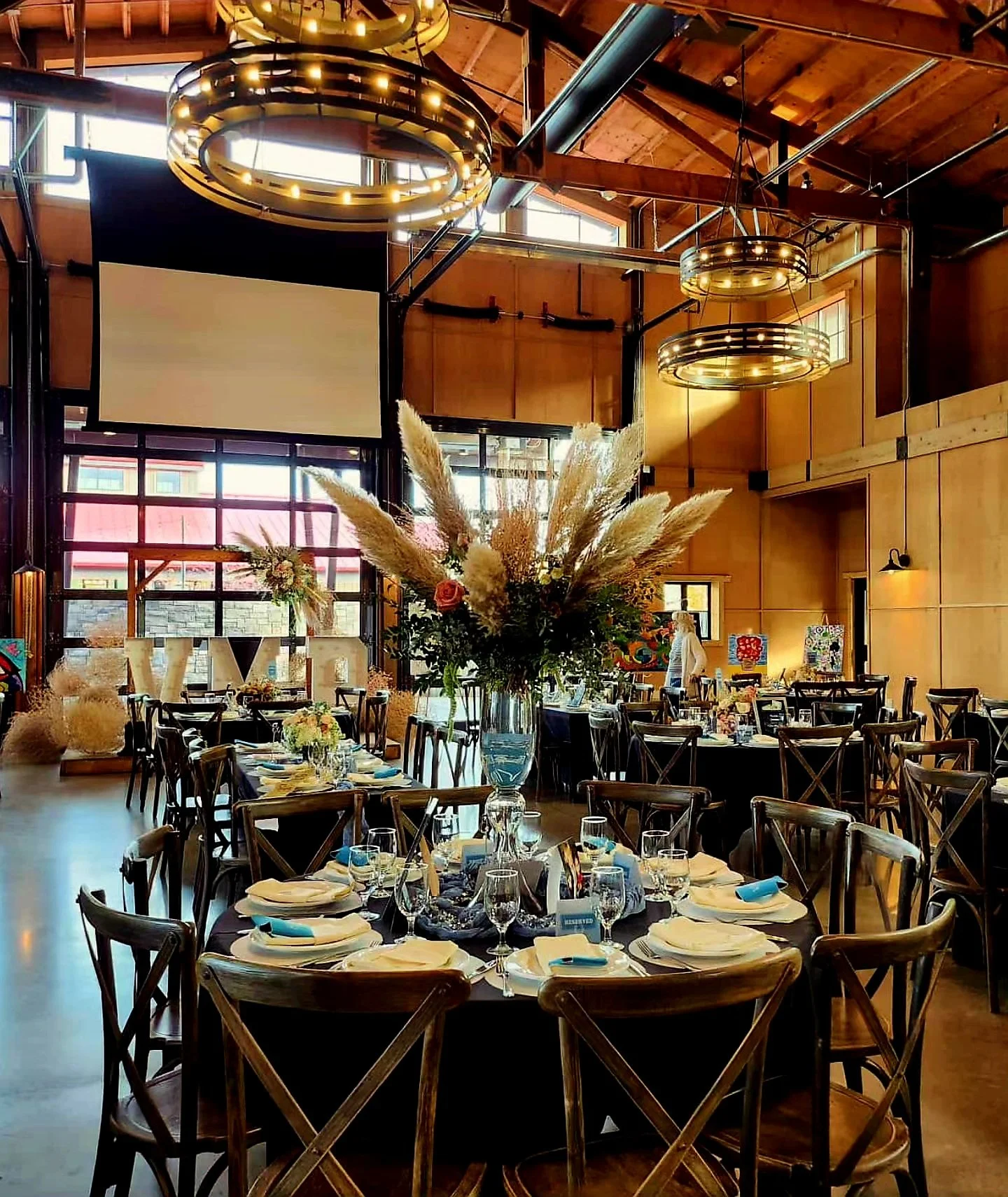 Elegant event space with round tables decorated with white napkins, glassware, and flower centerpieces, featuring a large floral arrangement on a central table, inside a wood-paneled room with high ceilings and modern circular light fixtures.