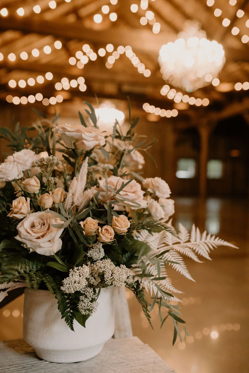 A floral arrangement with roses and greenery in a white vase, set in a warmly lit room with string lights and a chandelier in the background.