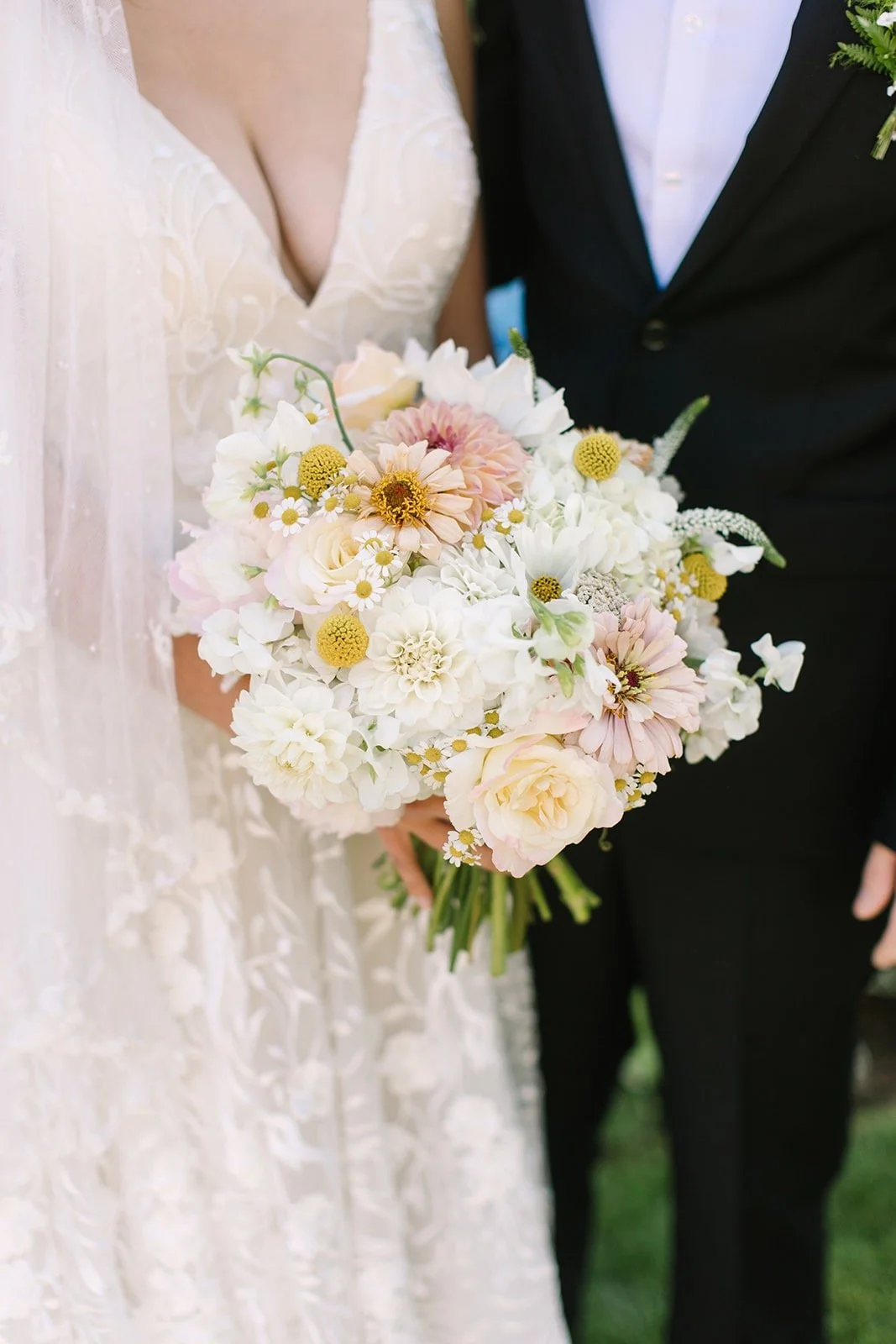 Close-up of a bride holding a bouquet of white and pastel pink flowers, standing next to a groom in a black tuxedo.