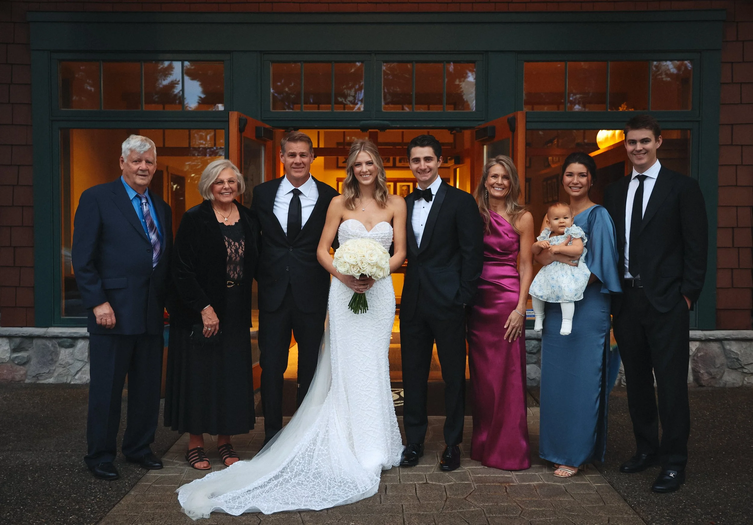 A group photo of nine people, including a bride and groom, standing in front of a building entrance. The bride is wearing a white wedding dress and holding a bouquet of white roses, while the groom is in a black tuxedo with a bow tie. The others are dressed in formal attire, with women in colorful dresses and men in suits. They are smiling and posing outdoors during the evening.