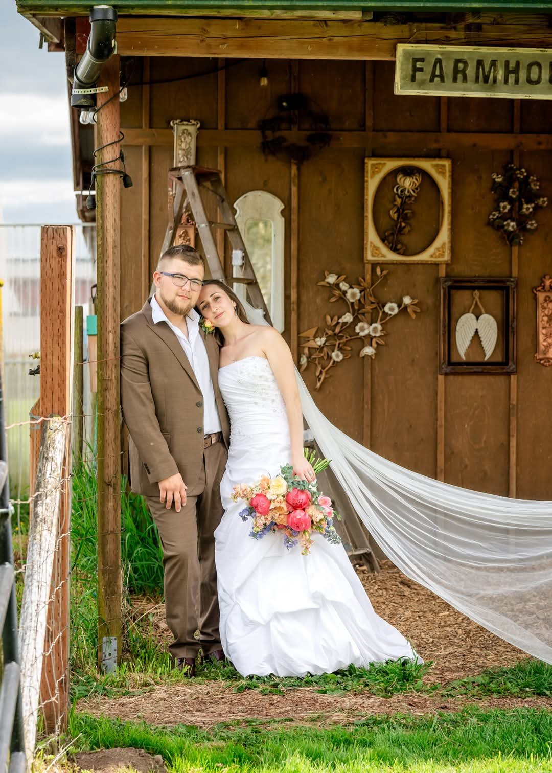 A bride and groom standing outdoors in front of a wooden structure decorated with art pieces and a sign that says 'FARMHOUSE.' The bride is in a white wedding gown holding a colorful bouquet of flowers, and the groom is in a brown suit with glasses.