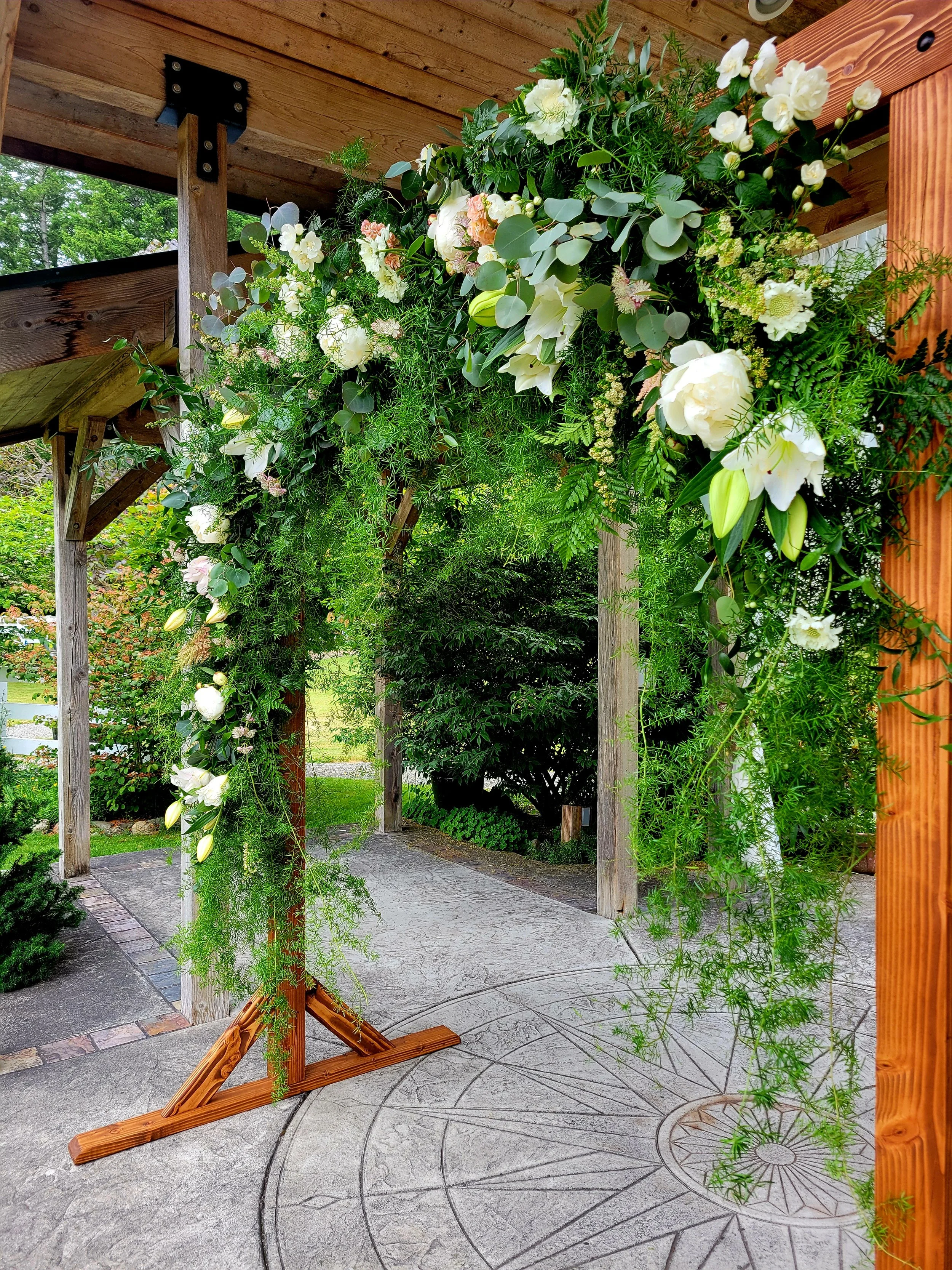 A decorative floral arch made of green leaves and white flowers, standing on a cement patio under a wooden roof. In the background, there are trees and garden plants.