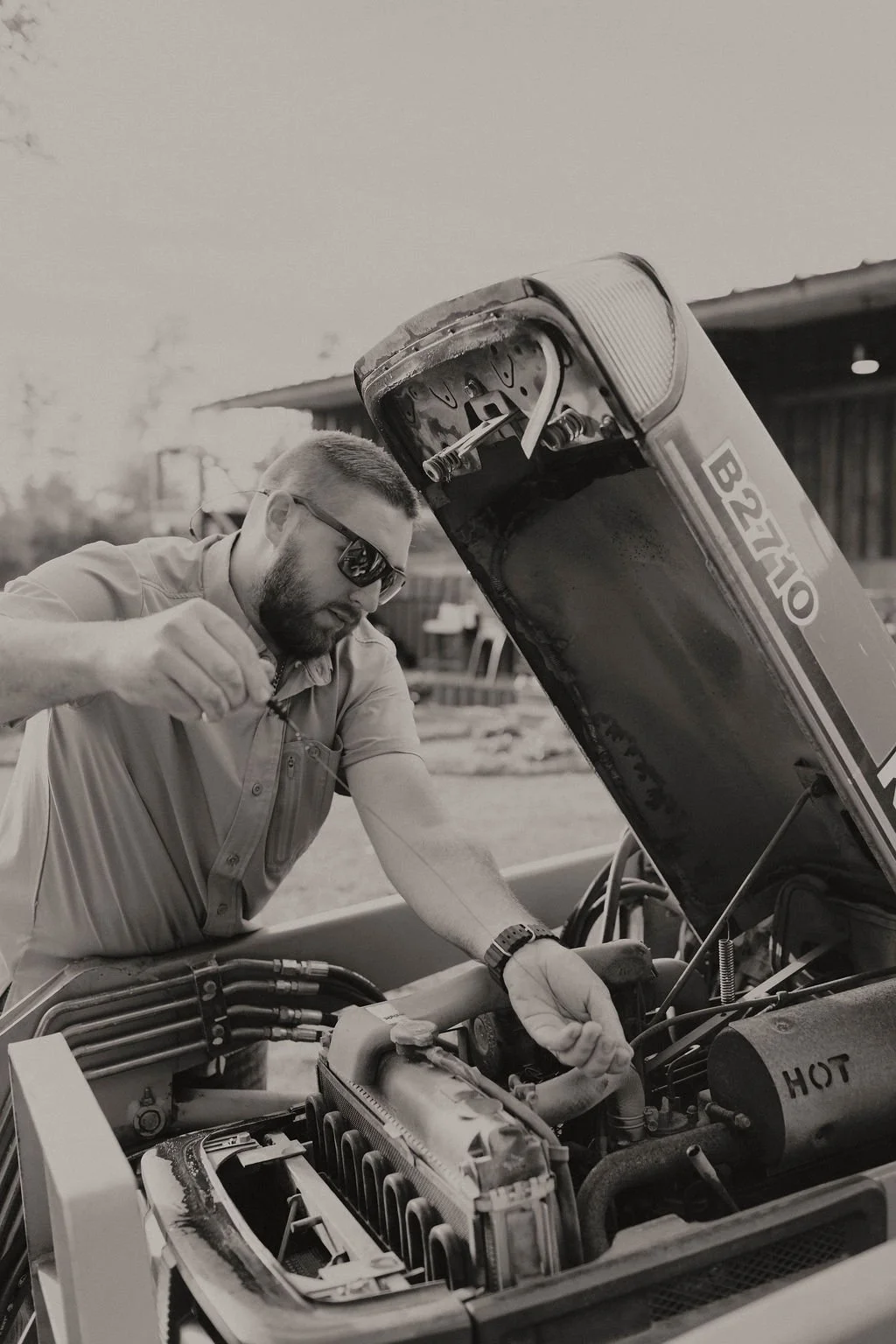 Mechanic wearing sunglasses repairs a vintage truck engine beneath the raised hood.