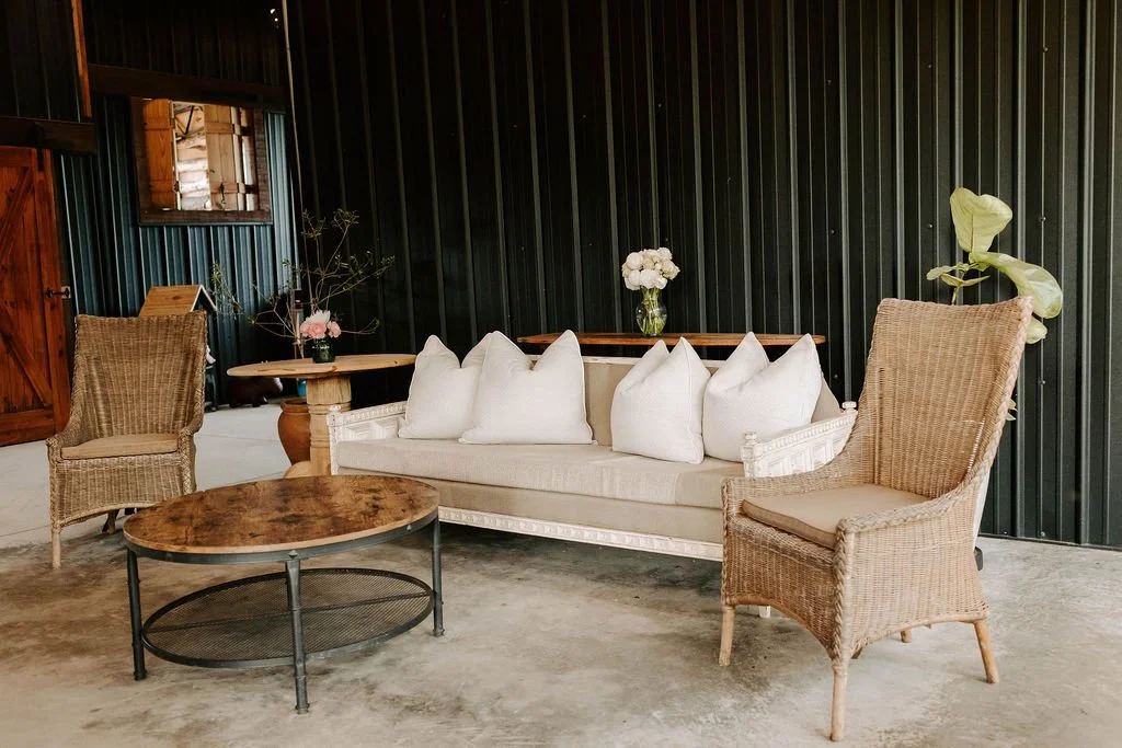 Rustic sitting area with a cream sofa, wicker armchairs, round wood coffee table, flowers, and dark corrugated metal walls