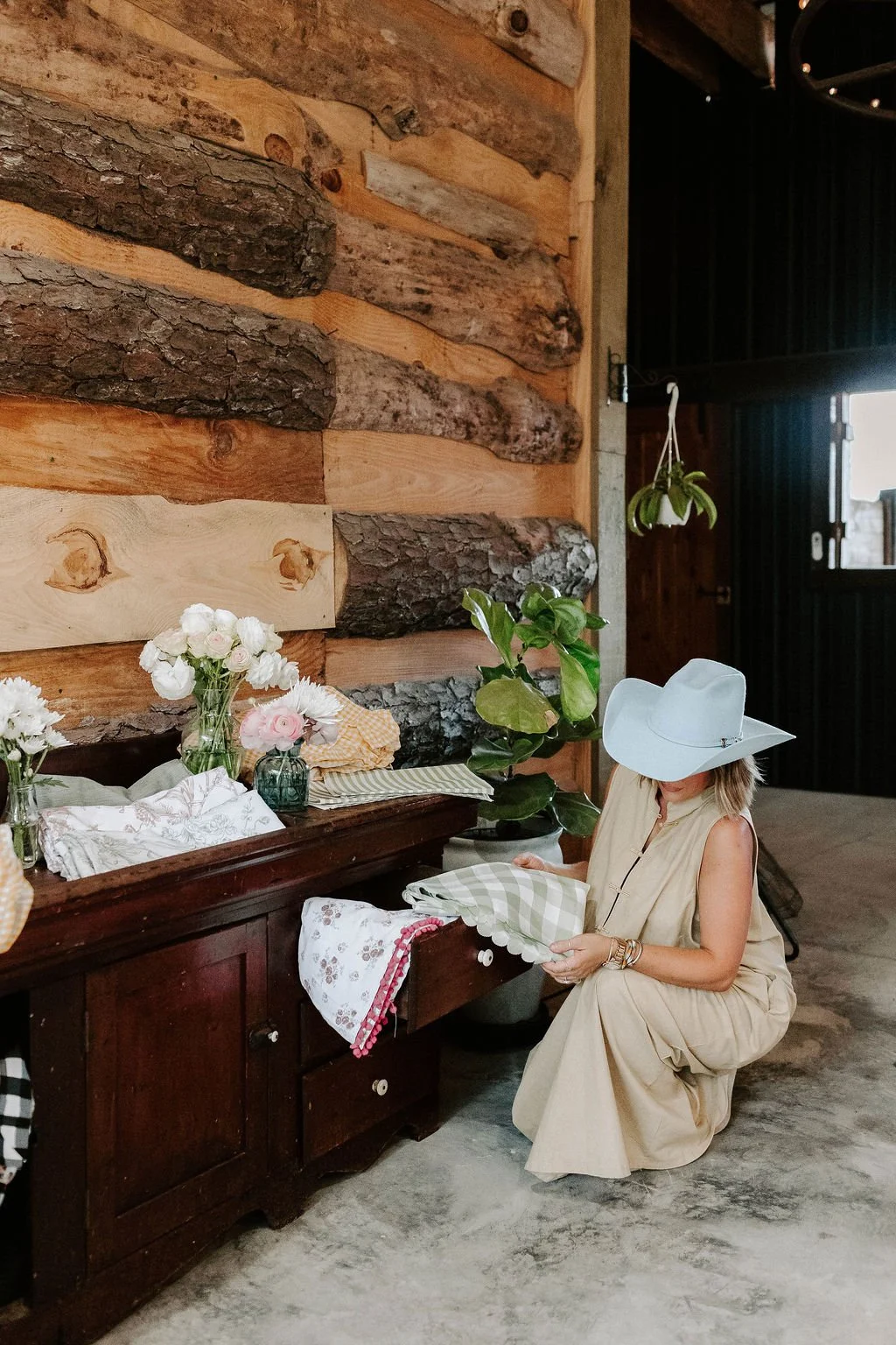 Woman in a white cowboy hat arranging floral fabrics beside a flower-decorated wooden cabinet in a rustic log cabin