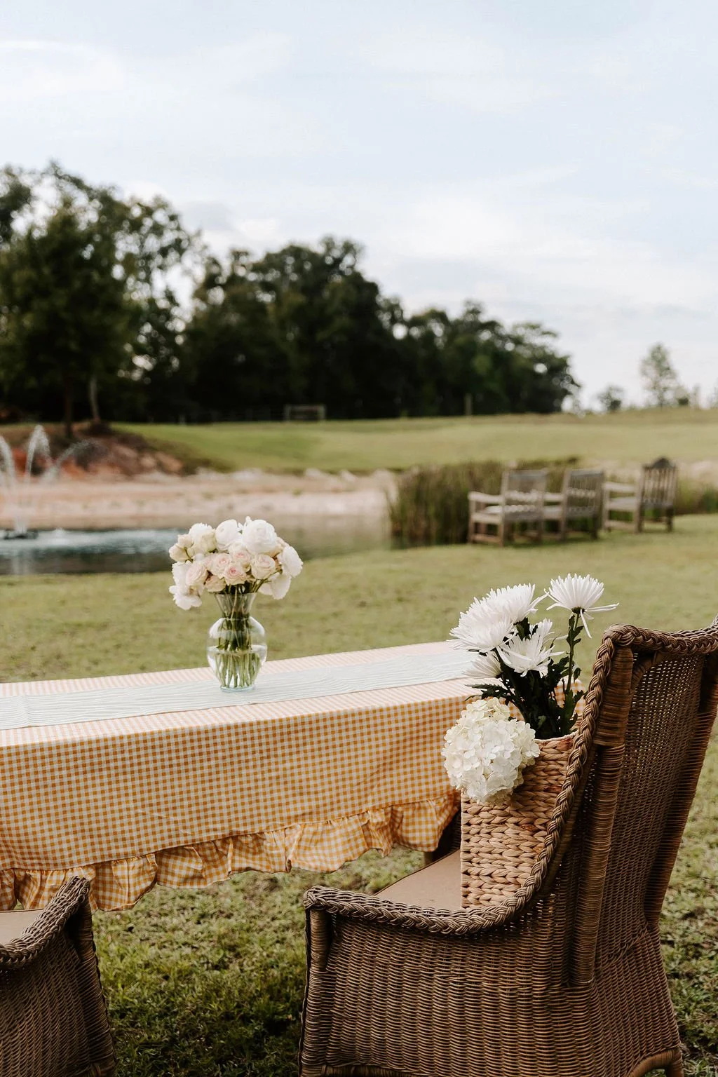 Outdoor table with orange gingham tablecloth, white floral arrangements, and wicker chairs beside a pond.