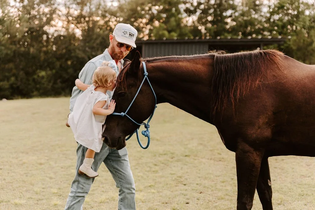 Man and young girl gently standing beside a brown horse in a grassy field.