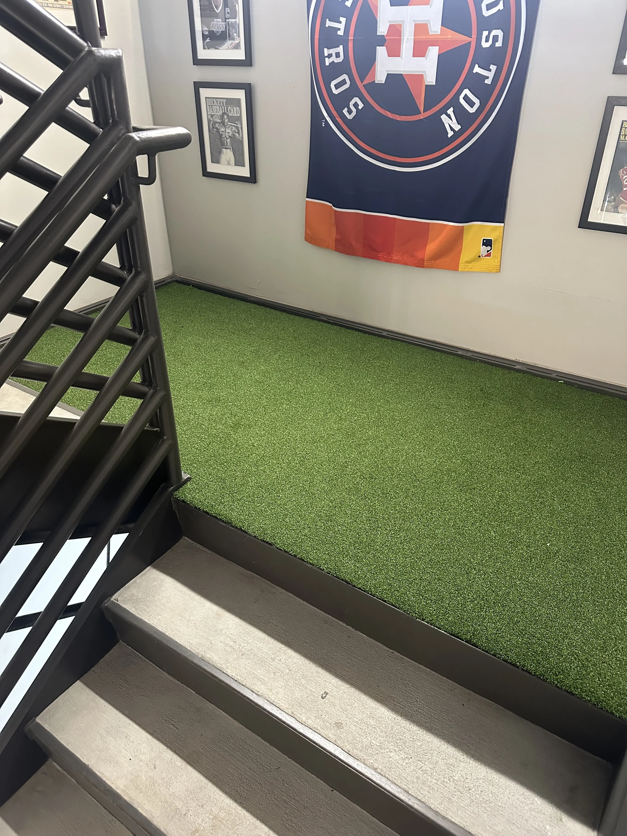 Staircase leading up to a small landing with a green artificial turf surface, framed photographs on the walls, and a large Houston Astros flag hanging on the wall.