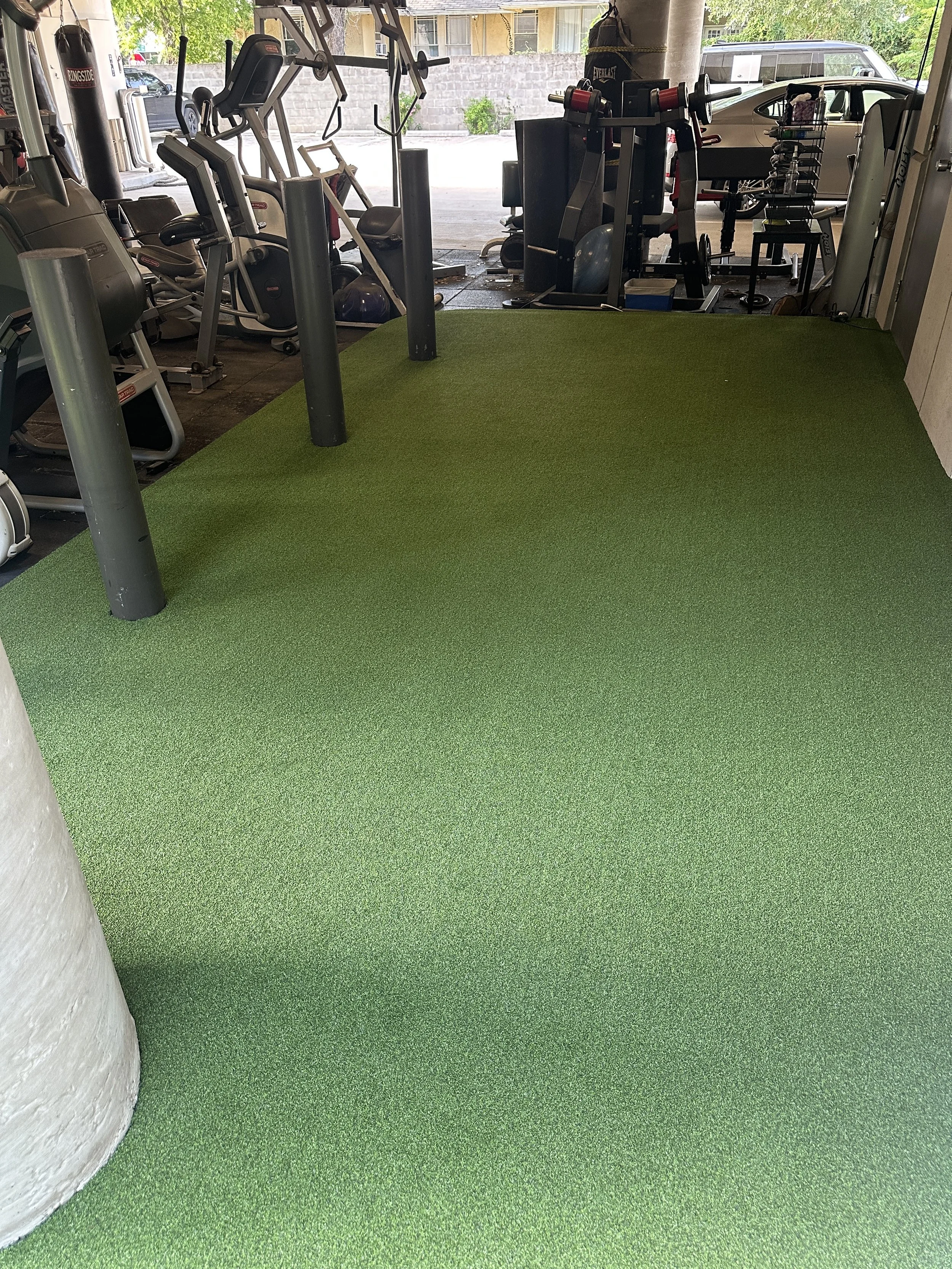 Indoor gym with green turf flooring laid by Houston's Horizon Turf & Greens, workout equipment like treadmills, ellipticals, and weight machines, with a view of parking lot outside.