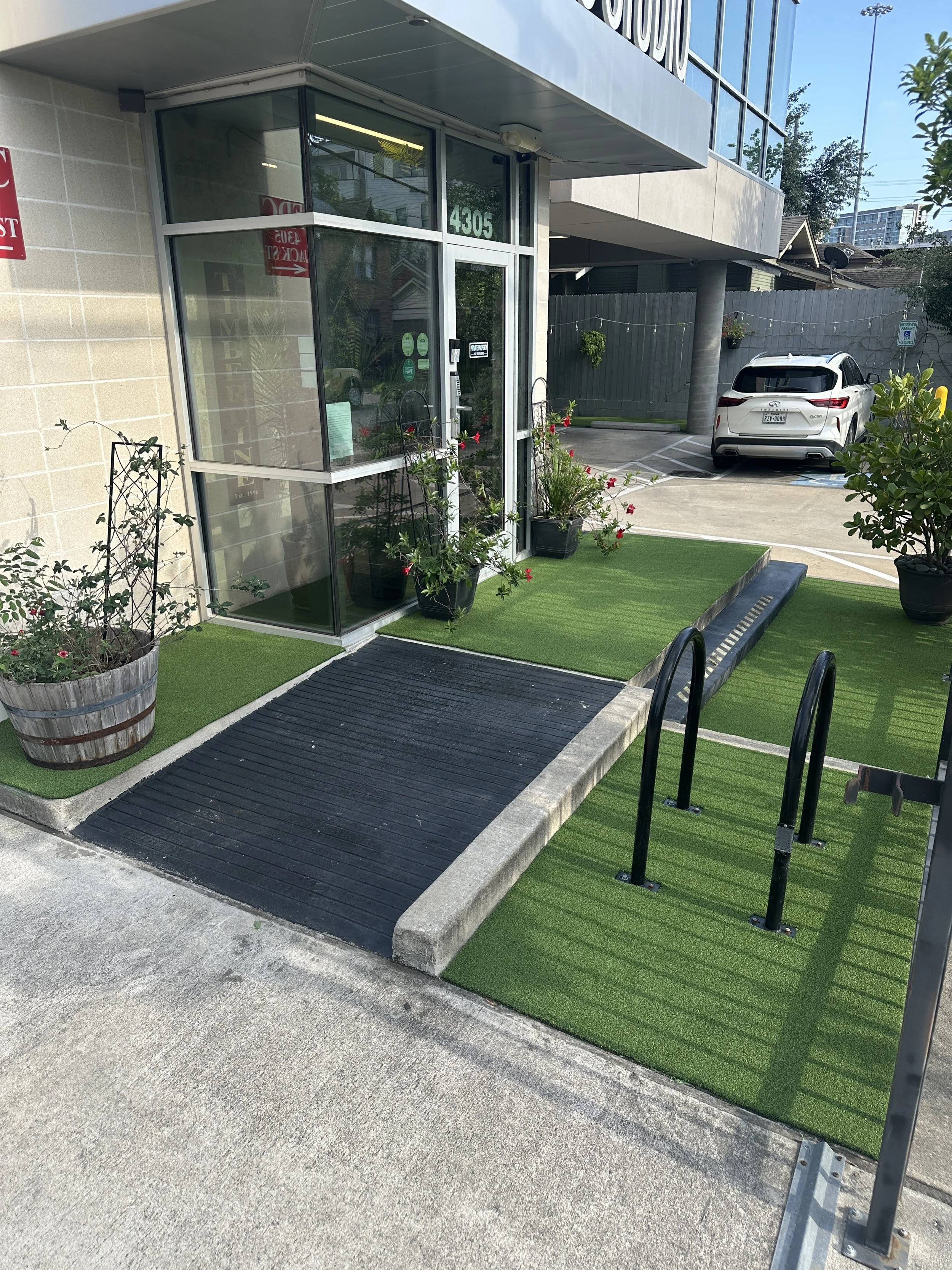 Entrance to a building with an accessible ramp, potted plants, and artificial grass at the entryway, located in a parking lot with a white SUV in the background.