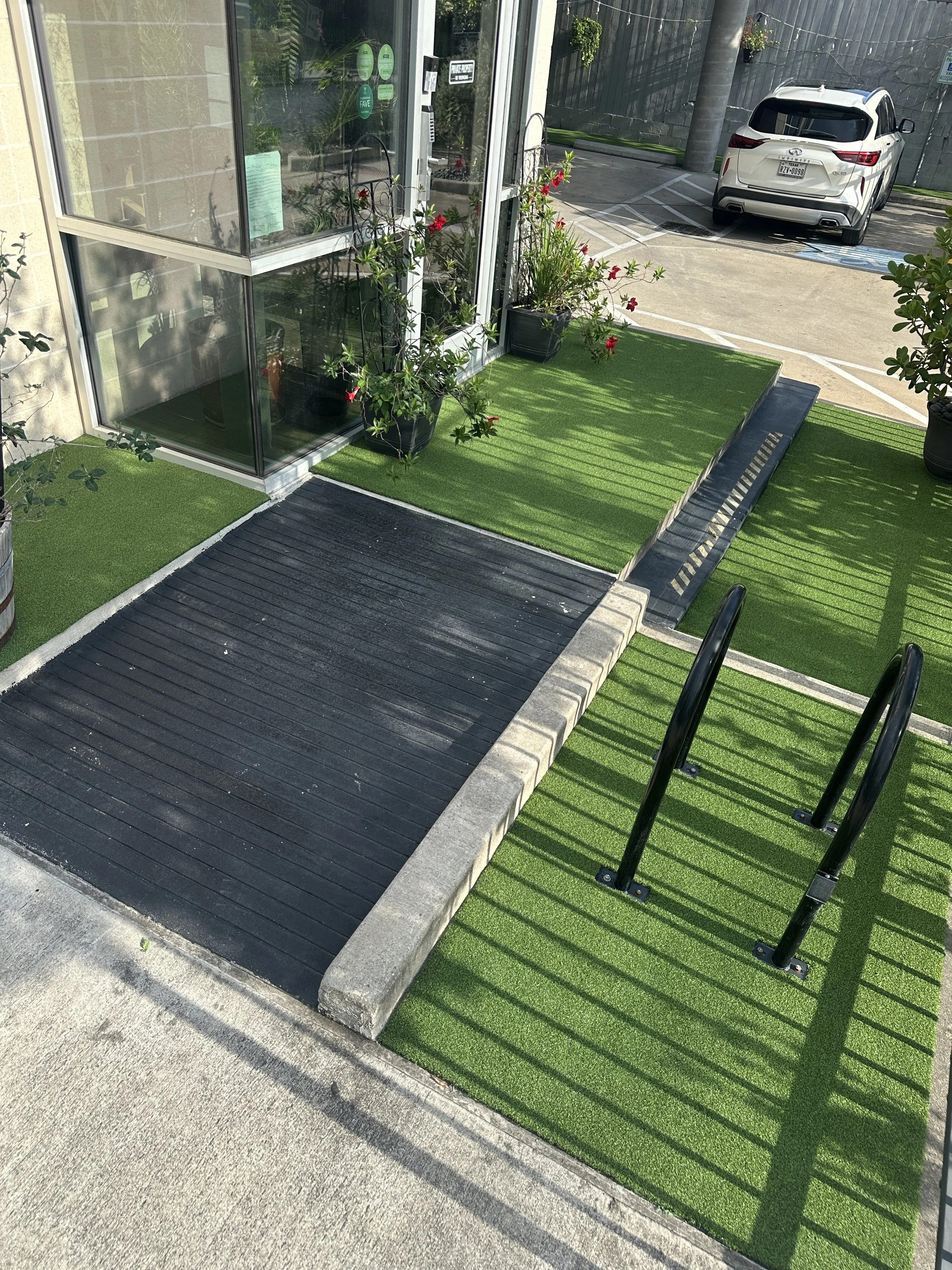 Outdoor entrance area with artificial grass, black rubber mat, potted plants, and a metal handrail. Turf laid by Horizon's Turf & Greens in Houston, TX.