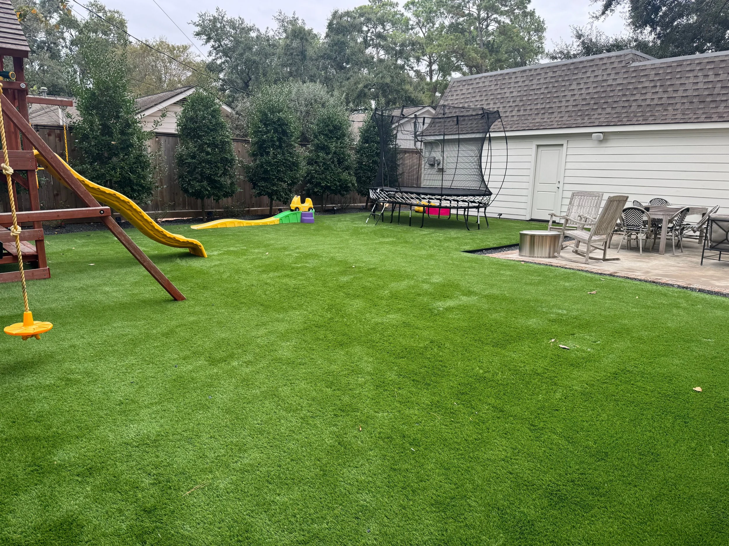 Backyard with artificial grass, yellow slide, trampoline, outdoor table and chairs, and a white shed.