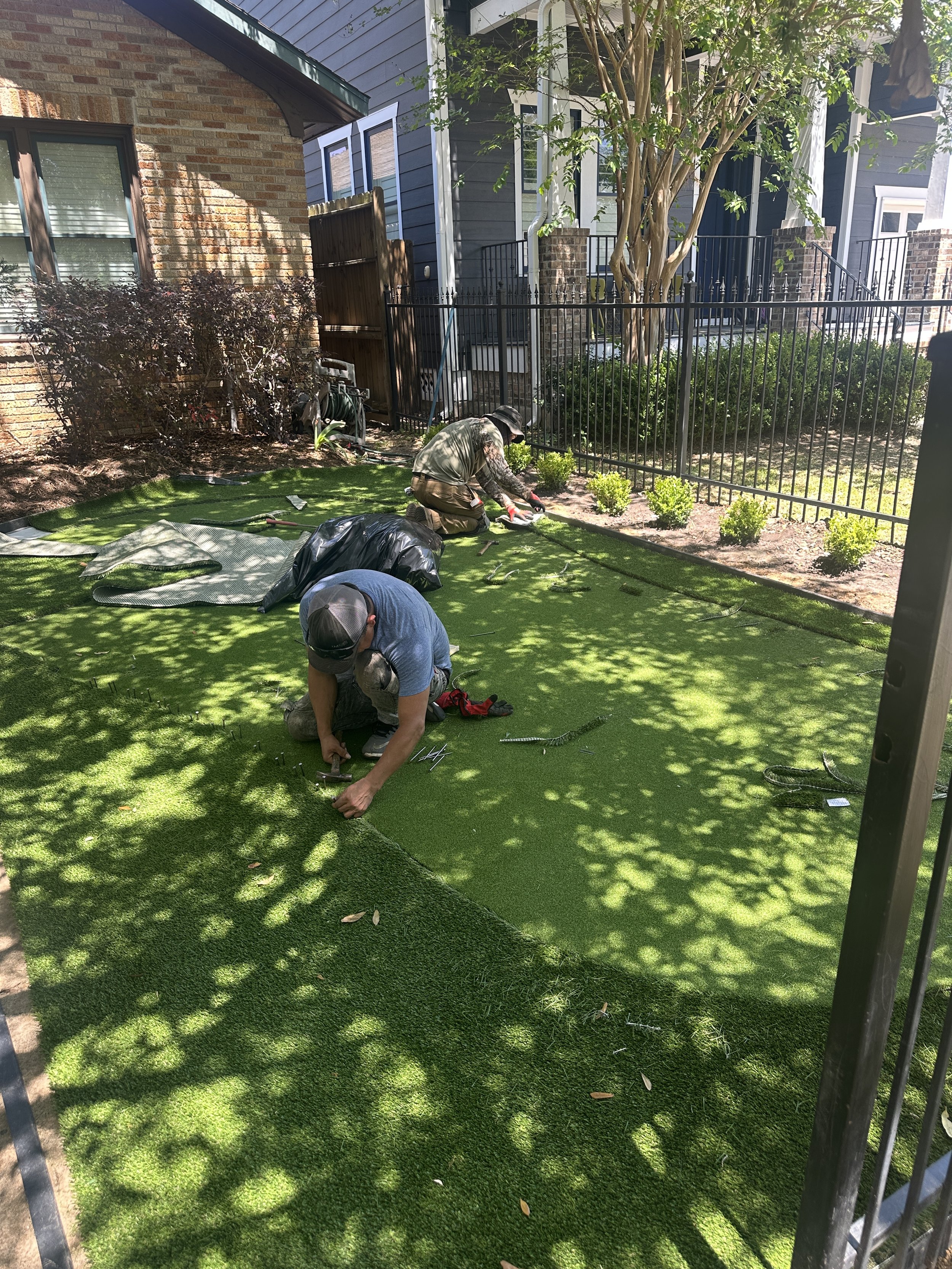 Two workers from Horizon Turf & Greens installing artificial grass on a Houston lawn, surrounded by a brick house and a blue house, with a tree providing shade.