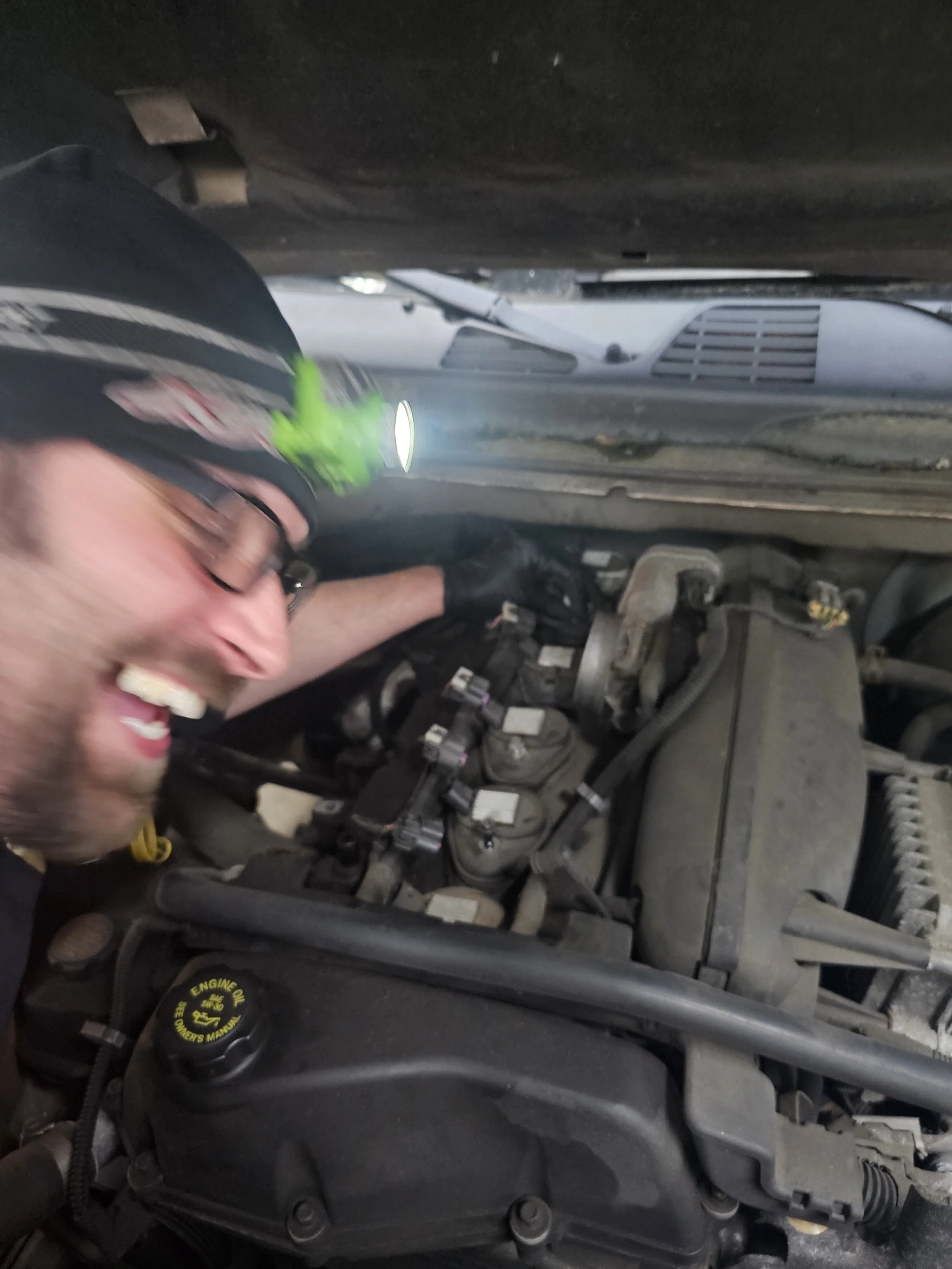 A man wearing safety glasses, gloves, and a cap with a headlamp, working under the hood of a car, inspecting or repairing the engine.