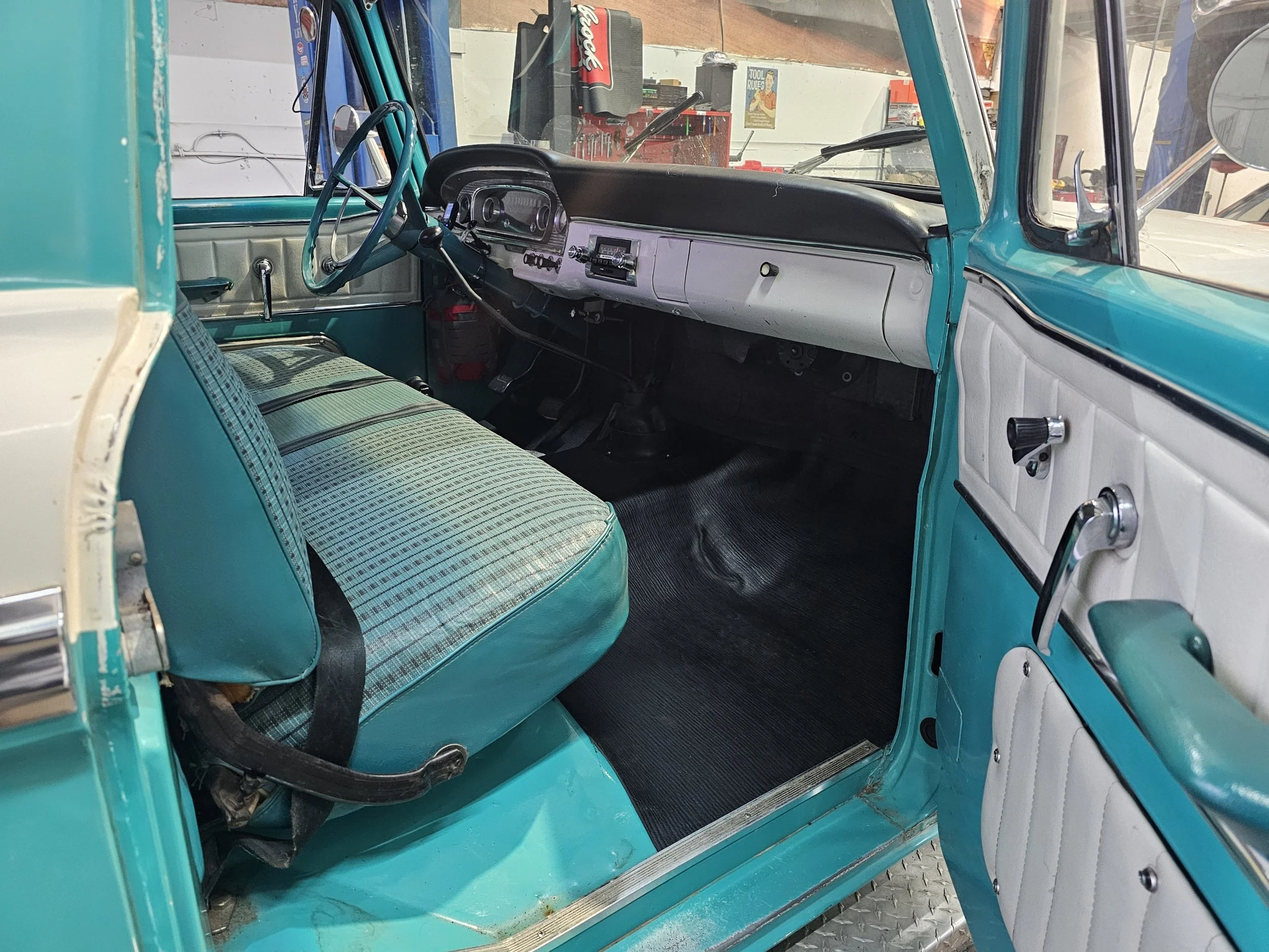 Interior of a vintage turquoise and white truck showing a plaid cloth seat, steering wheel, dashboard, and manual window cranks.