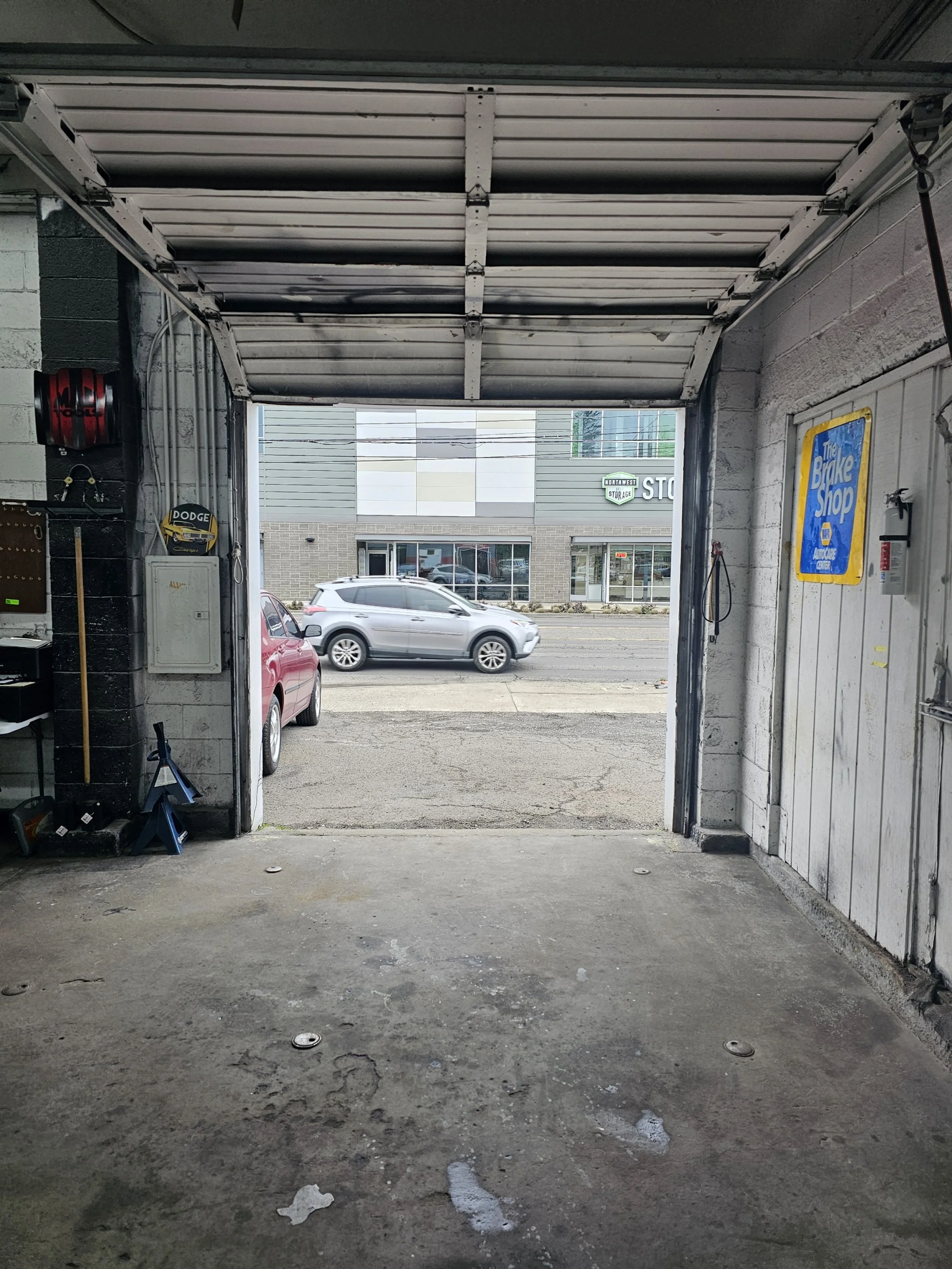 View from inside an open garage looking out onto the street with a red car parked on the left and a silver SUV driving by. Buildings and storefronts are visible across the street.