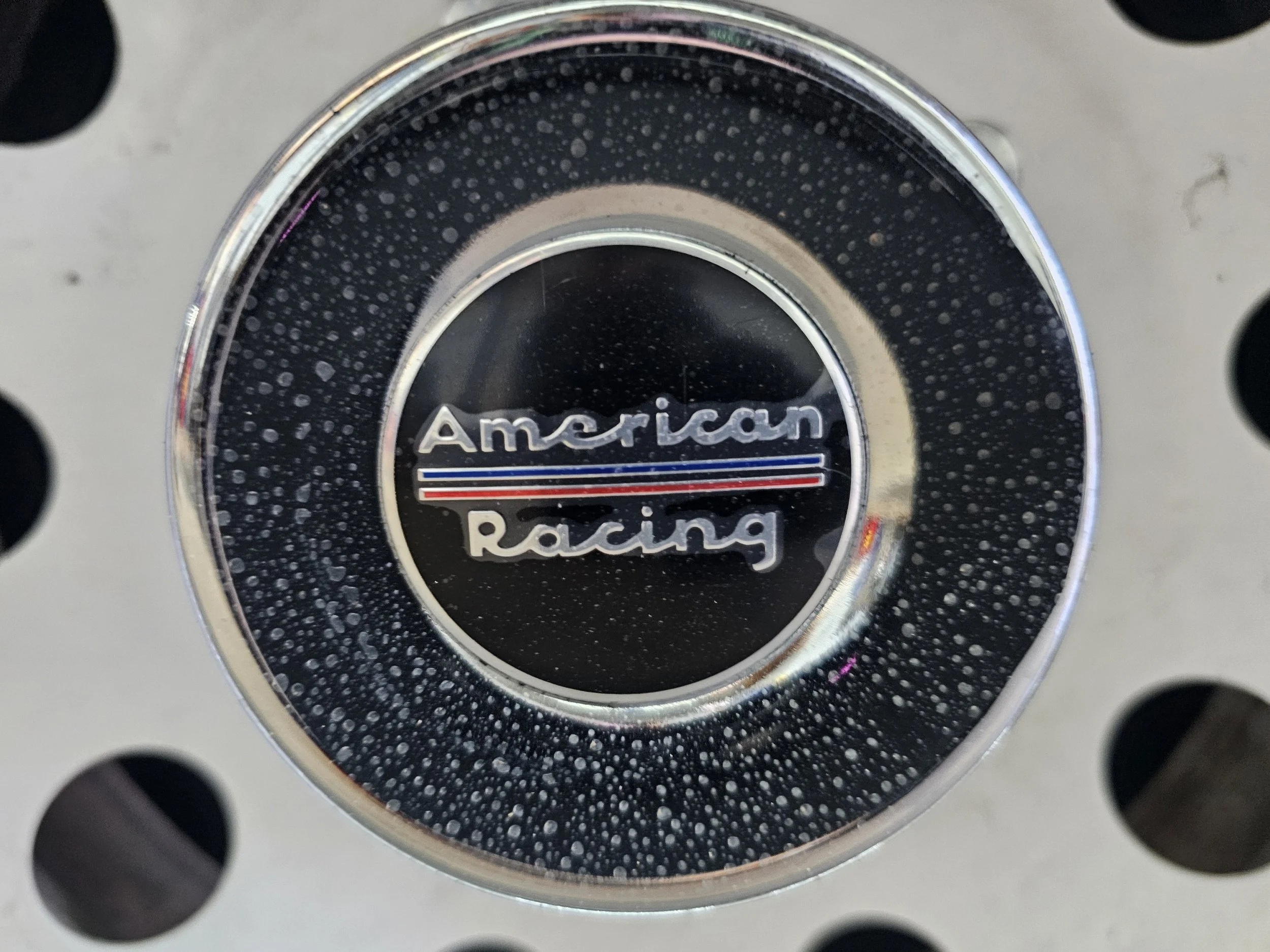 A black circle with the words 'American Racing' in silver, surrounded by a silver ring with water droplets, set against a perforated metal background.