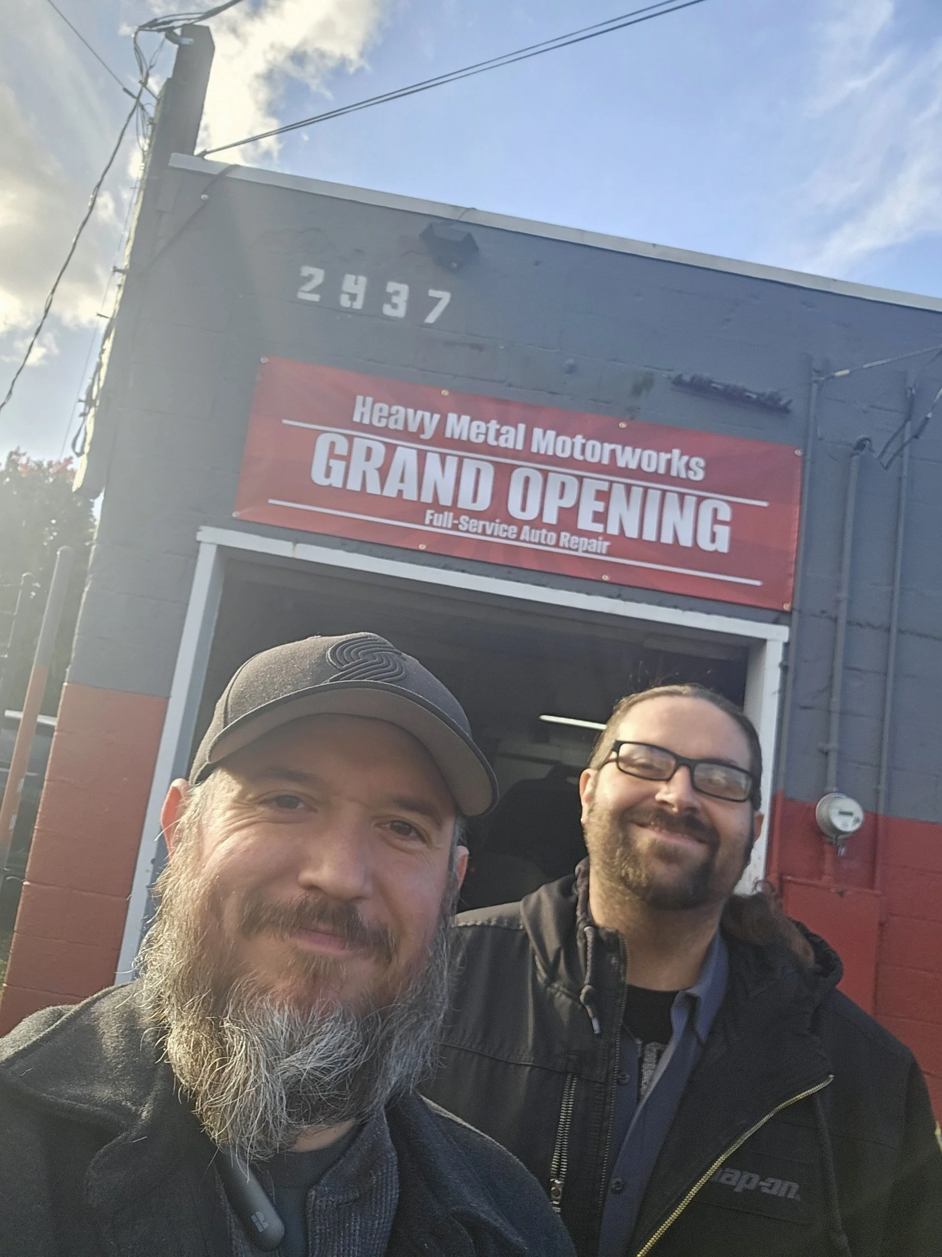Two men smiling in front of a gray building with a red sign that reads 'Heavy Metal Motorworks GRAND OPENING Full-Service Auto Repair'. One man has a gray beard and is wearing a gray baseball cap, the other has dark hair, glasses, and a dark jacket.