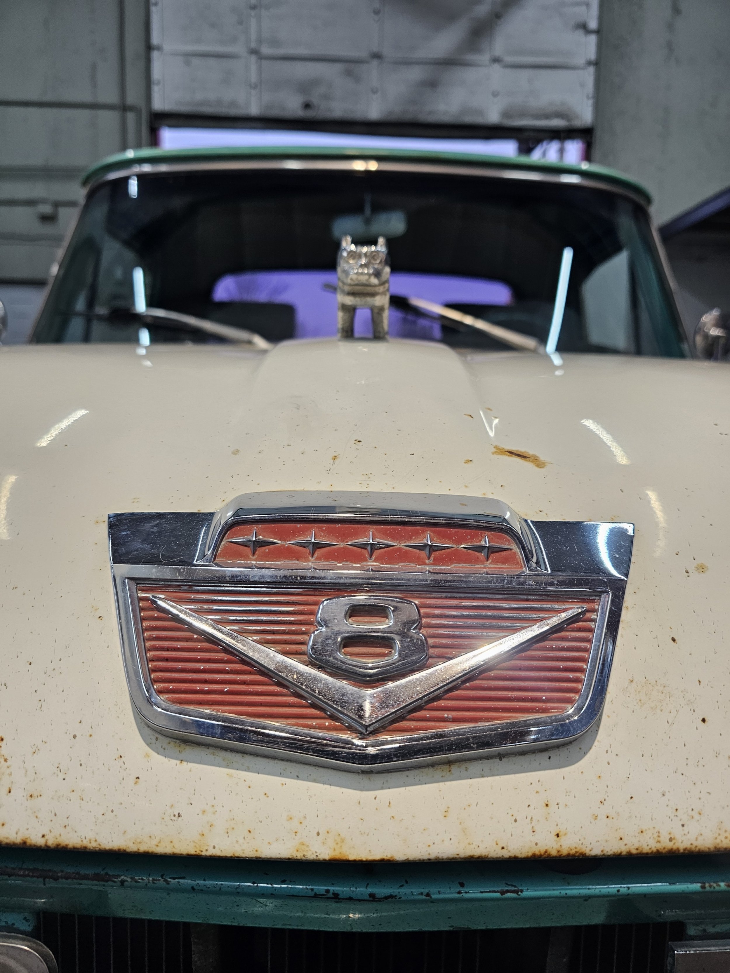 Front view of a vintage cream-colored car with a rusted hood ornament and an emblem featuring the number 8 and a V-shaped design.