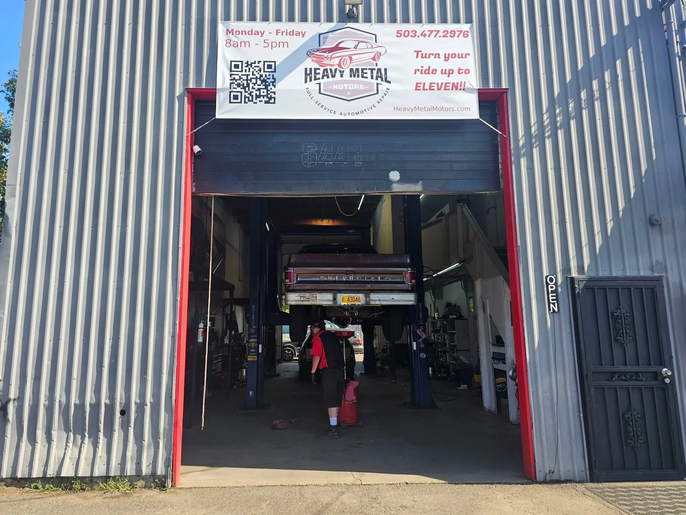 Auto repair shop with a truck lifted in the air inside the garage. A mechanic or worker is behind the truck. A sign above the garage shows the business name "Heavy Metal Motors" and hours of operation.