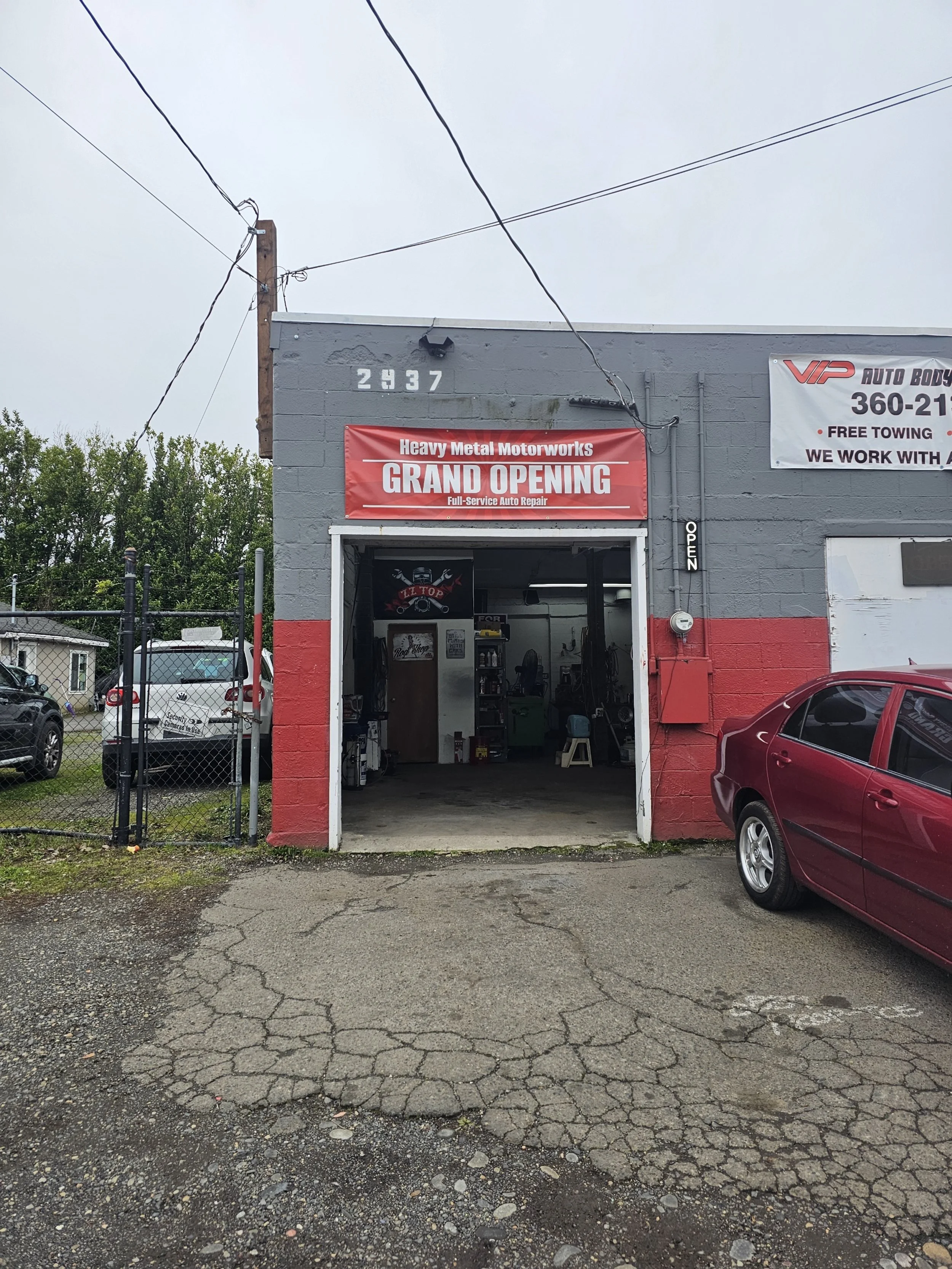Exterior of an auto repair shop with a red banner that says 'Heavy Metal Motorworks Grand Opening,' with a partially open garage door revealing the interior of the workshop, a red car parked in front, and a black fence on the left side.