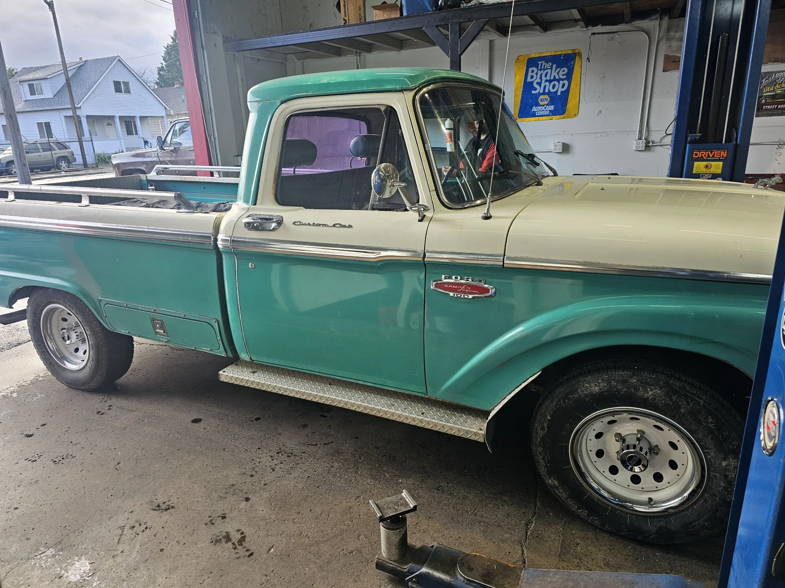 A vintage green and cream Ford pickup truck inside an auto repair shop. The truck has a step side with a chrome side mirror and a camper badge on the door. The shop has a blue sign that reads 'The Brake Shop' and various tools and equipment around.