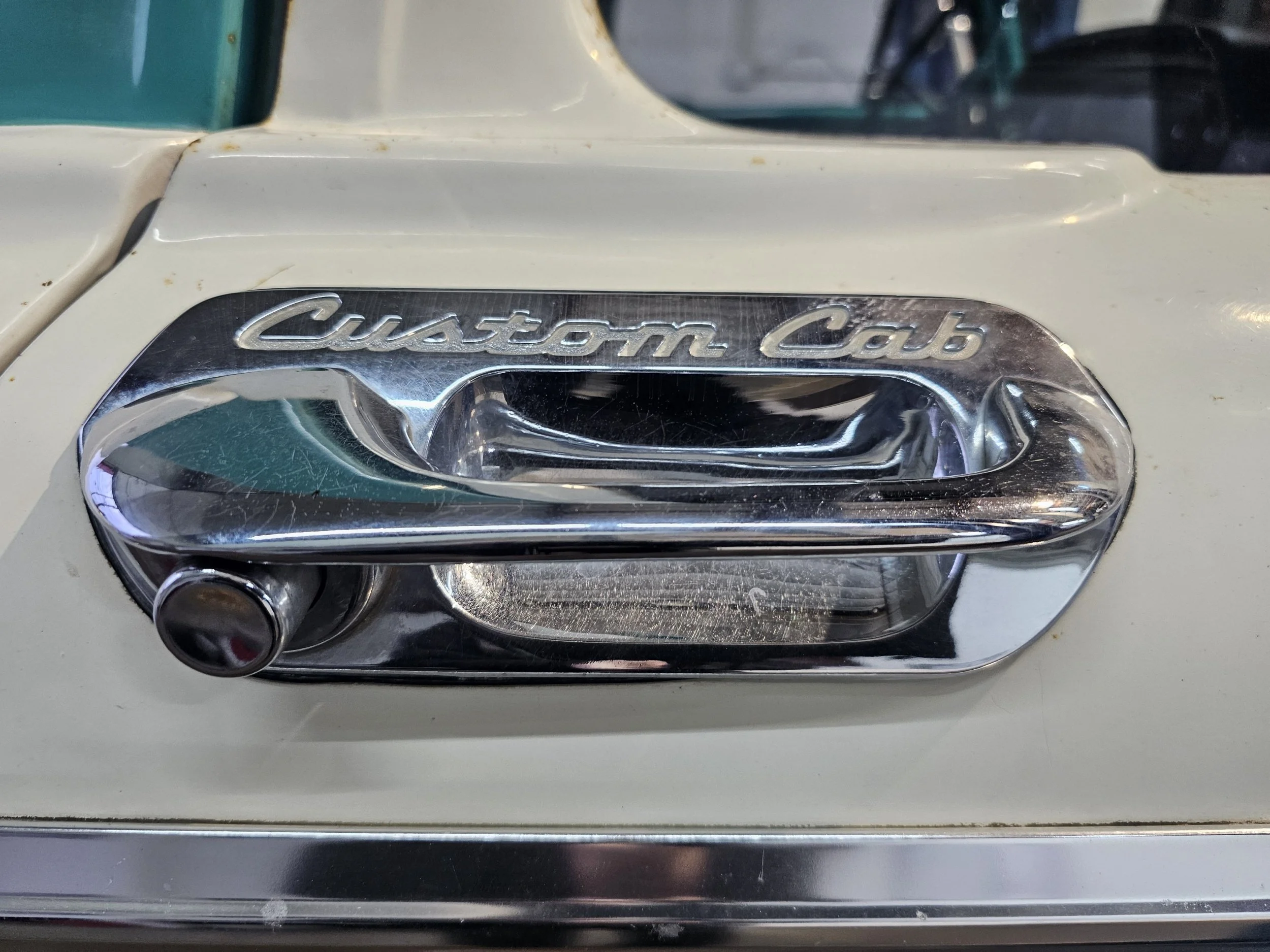 Close-up of a vintage car door handle with the inscription 'Custom Cab' in chrome lettering, attached to a cream-colored vehicle.