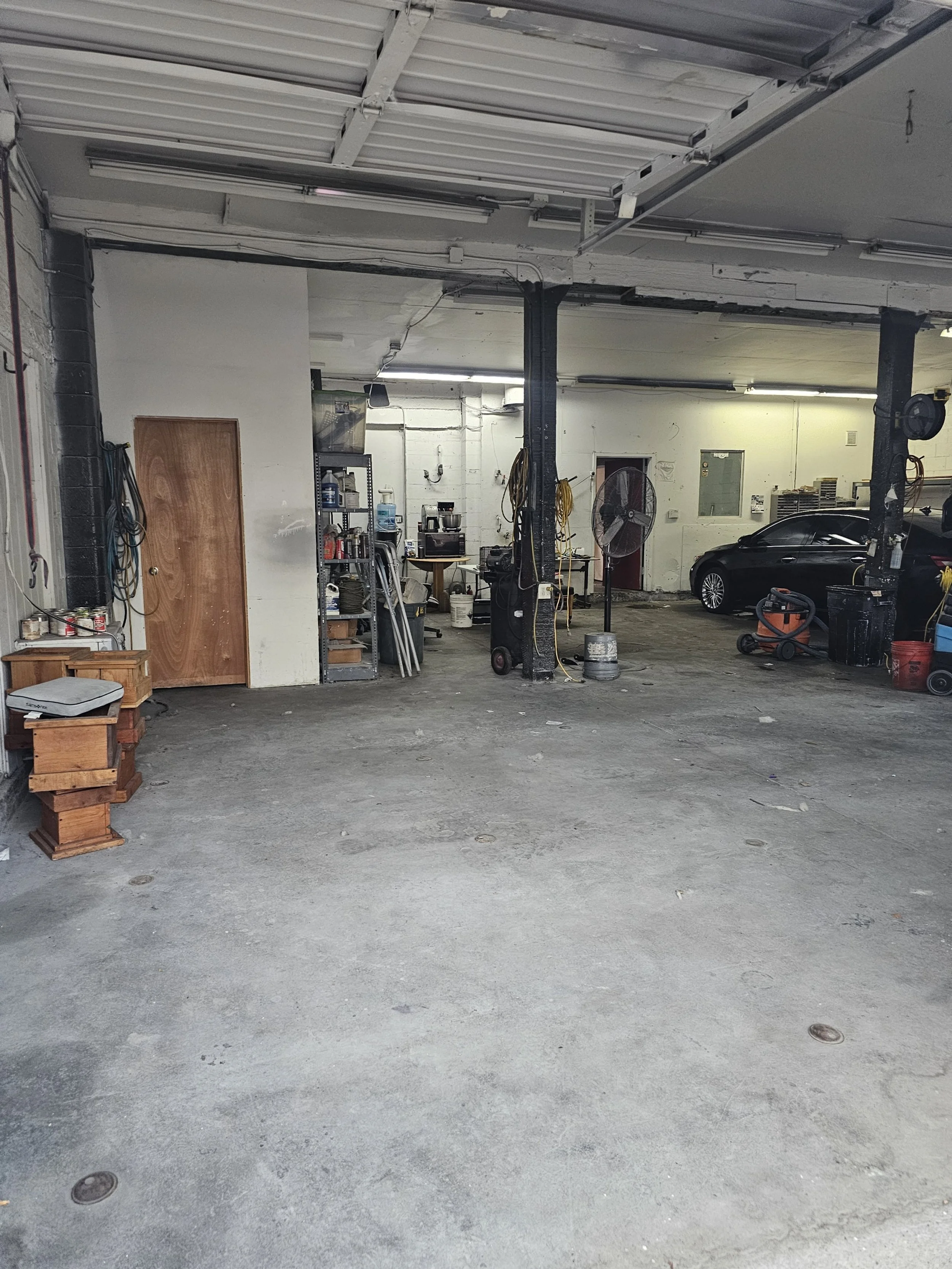 Indoor garage with concrete floor, black car, tools, shelves, and a large fan.