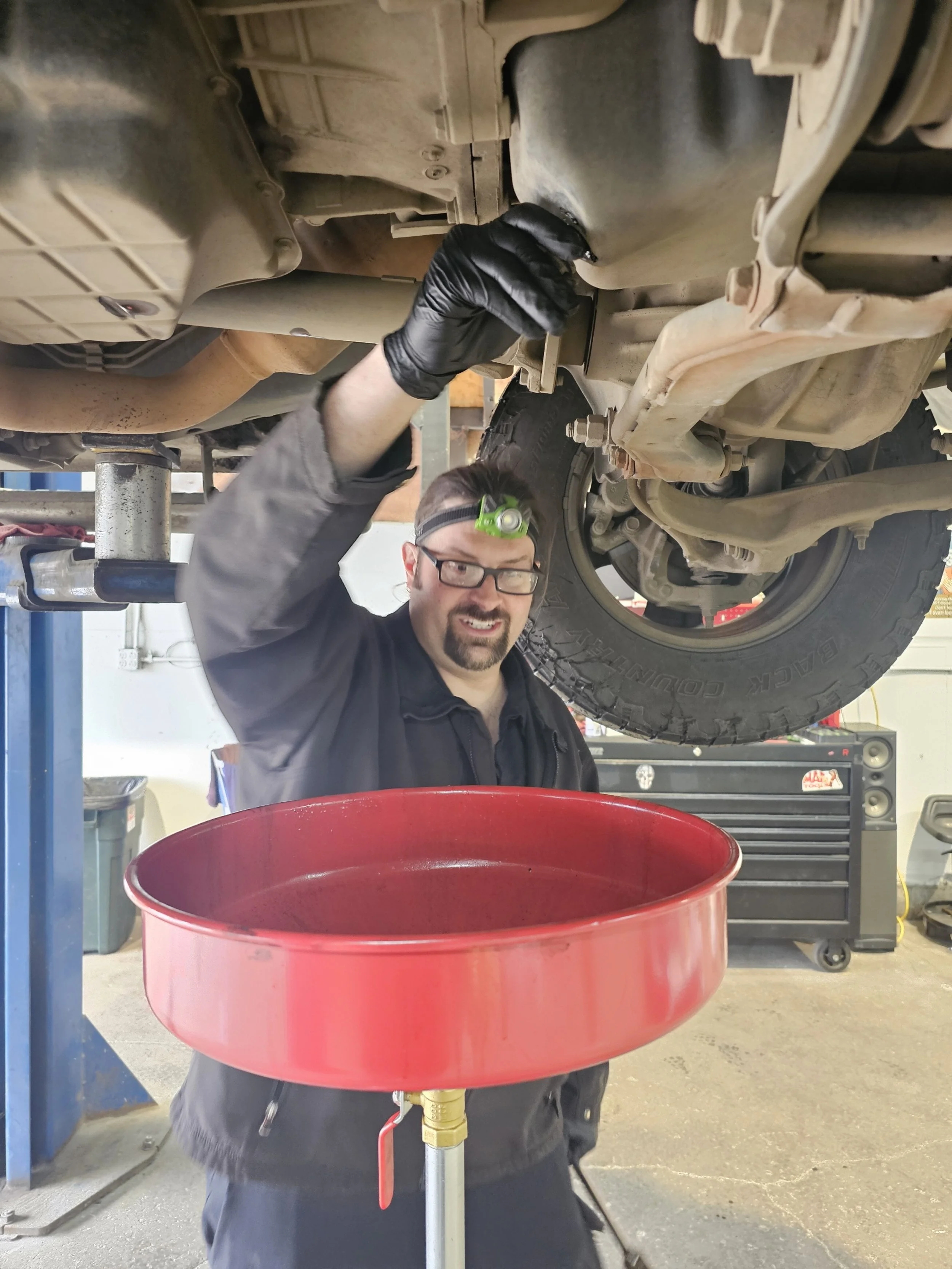 A mechanic working underneath a vehicle elevated on a lift, with a red oil drain pan underneath, in a garage or workshop.