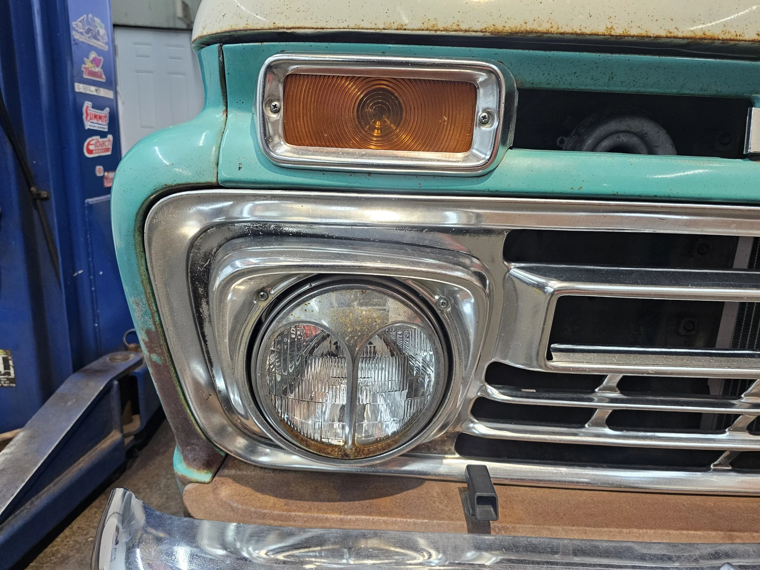 Close-up of the front of a vintage turquoise and chrome vehicle, showing a rectangular amber turn signal light and a round headlight beneath a chrome grille.