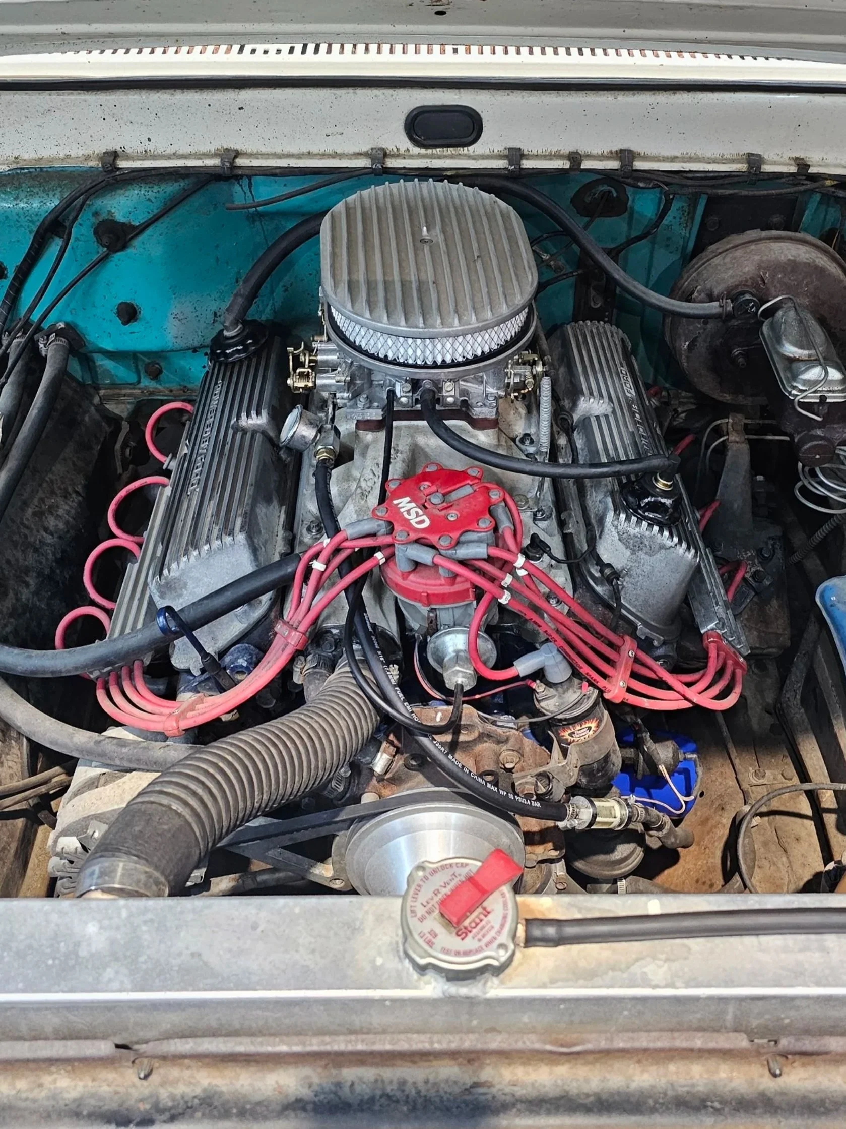 Inside a car engine bay, showing a V8 engine with red ignition wires, a silver air filter on top, and various hoses and components.
