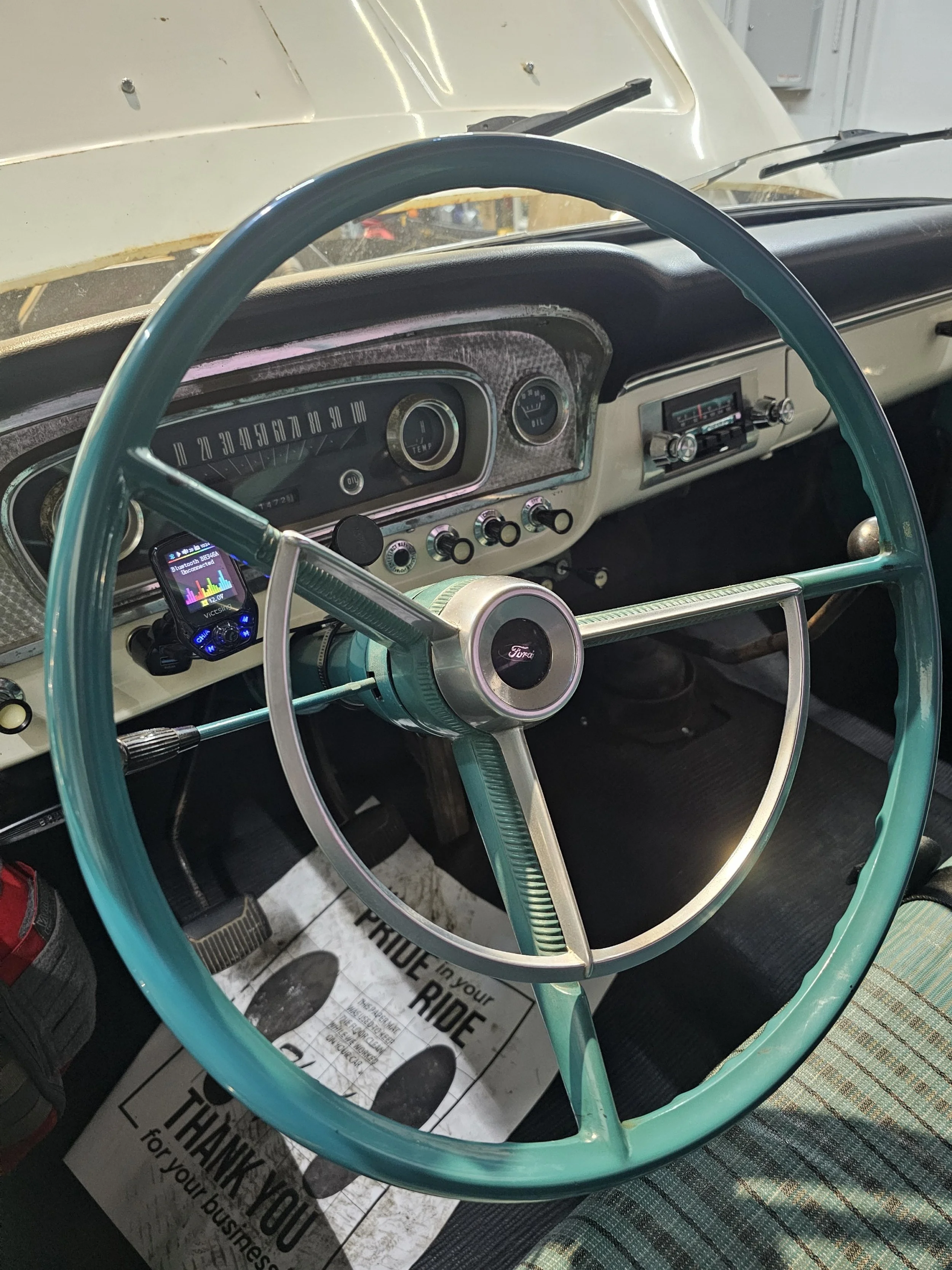 Interior of a vintage Ford truck showing a teal steering wheel, dashboard with speedometer, oil gauge, and vintage radio, with a smartphone mounted on the dashboard displaying music.