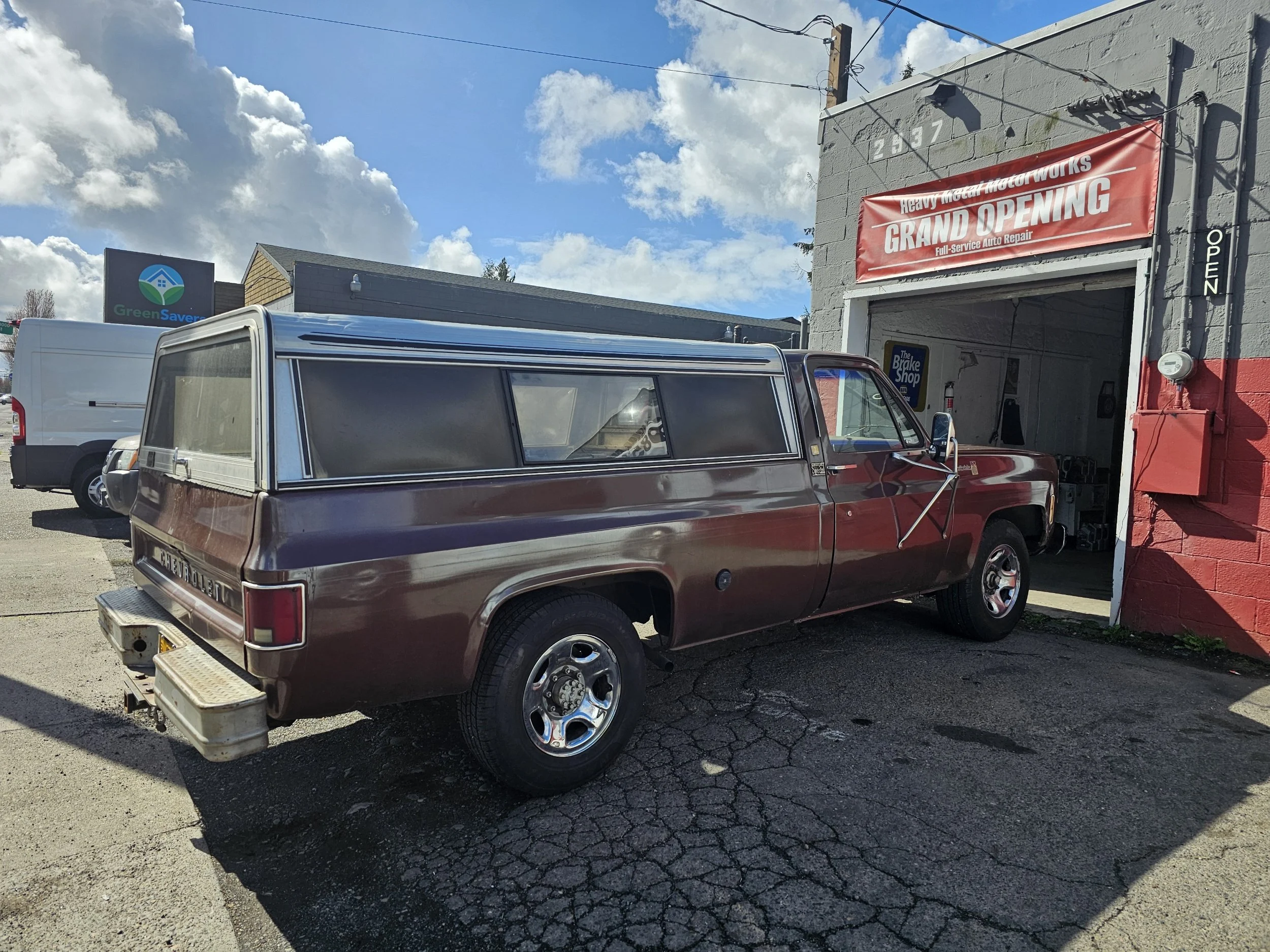 A vintage brown Chevrolet pickup truck with a matching camper shell parked outside an auto repair shop with a red sign that reads 'Grand Opening.' The shop has an open garage door, and a white van is parked nearby under a blue sky with scattered clou