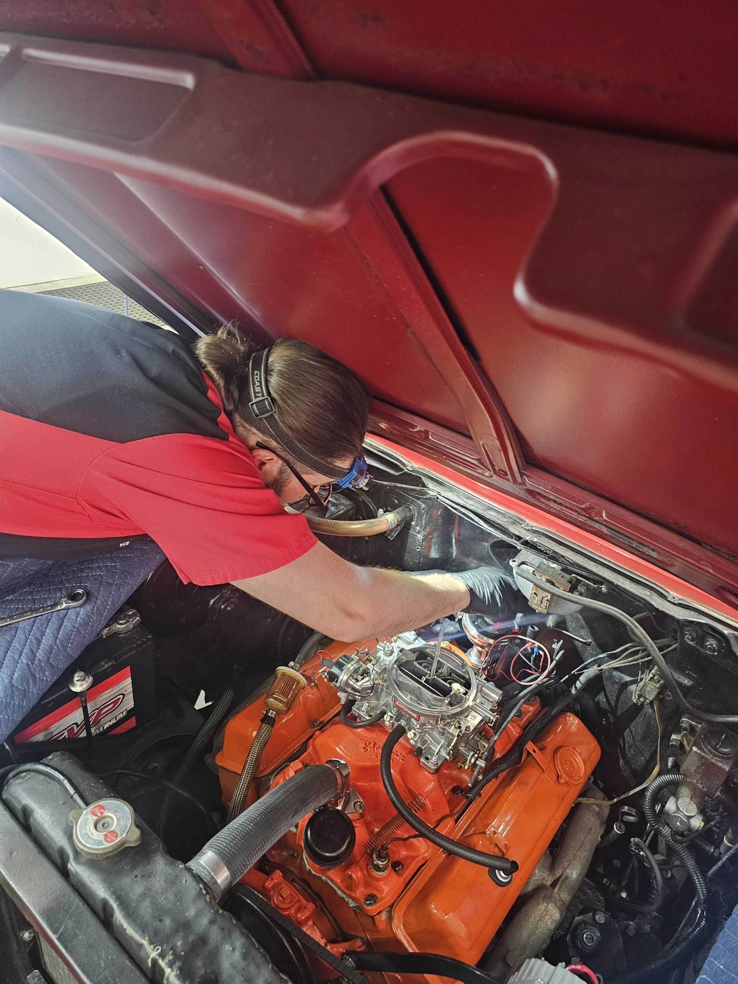 A person wearing glasses and a red and black shirt working on a car engine in a garage.