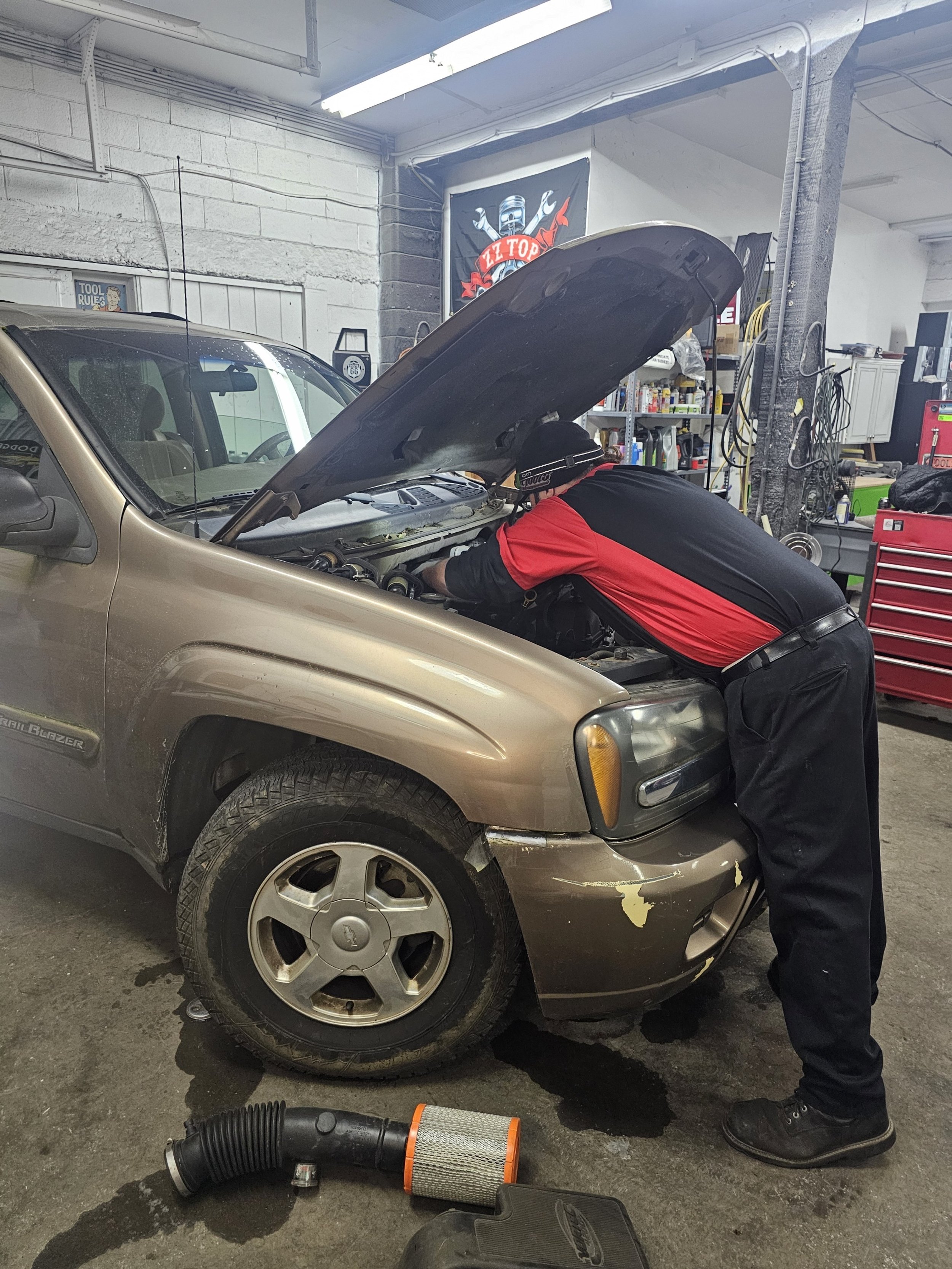 A mechanic working under the hood of a gold Chevrolet TrailBlazer in an auto repair shop.