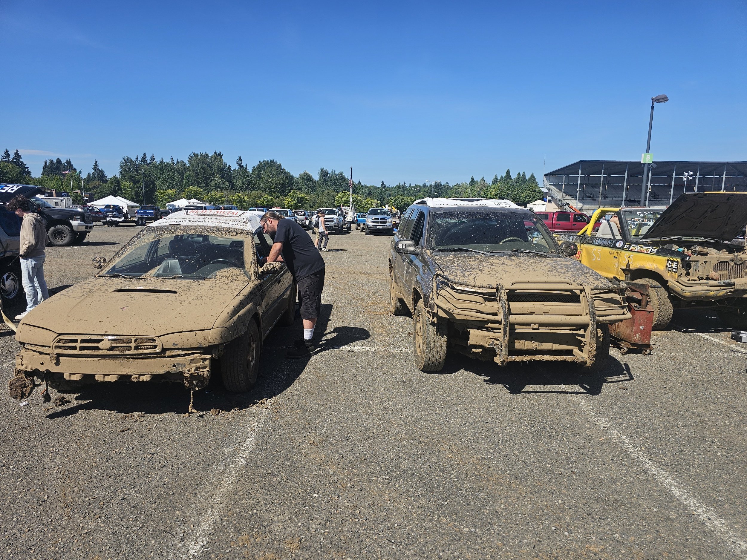 Two muddy, damaged race cars in a parking lot, with people inspecting them and a crowd of vehicles in the background.