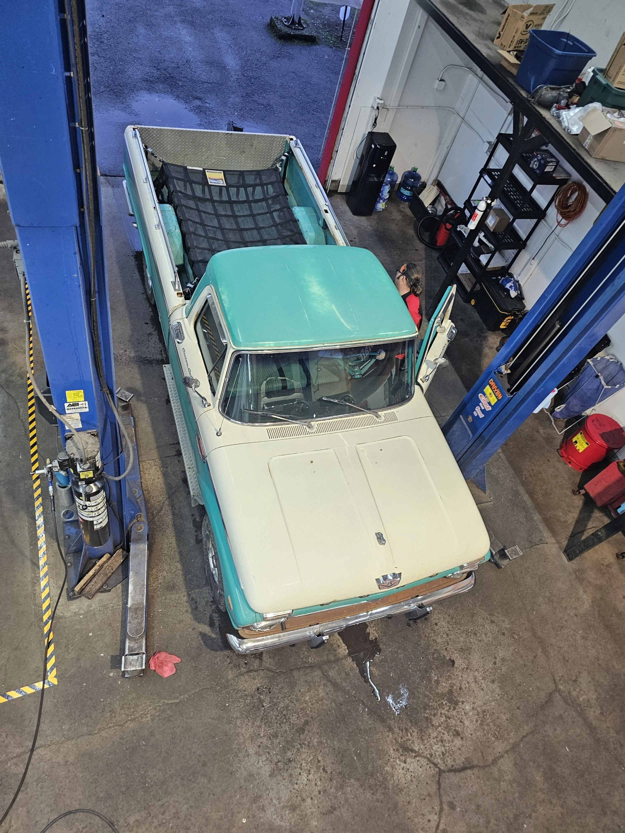 An overhead view of a vintage pickup truck inside a garage. The truck is cream-colored with a teal roof and tailgate, and it is positioned on a hydraulic lift. A person is working on the driver side, near the open door. The garage has shelves with tools and supplies, water bottles, a water cooler, and boxes.